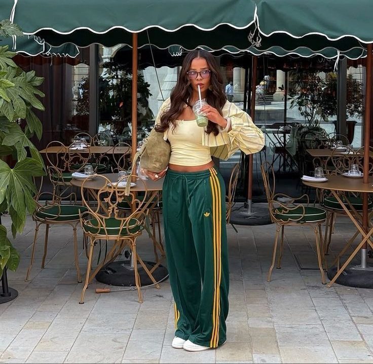 Mulher jovem com cabelos longos e &oacute;culos, vestindo top amarelo claro, jaqueta listrada e cal&ccedil;a verde Adidas com listras amarelas, segurando uma bebida verde com canudo e uma bolsa verde, em frente a um caf&eacute; com mesas e guarda-s&oacute;is verdes