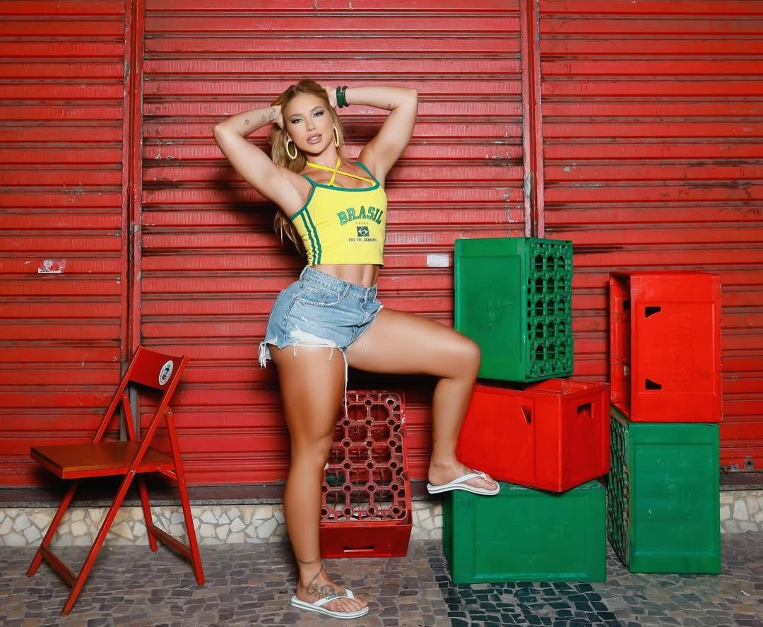Mulher loira, com top amarelo e verde escrito Brasil, shorts jeans e chinelos brancos, posa com uma perna sobre caixas de pl&aacute;stico vermelhas e verdes, em frente a uma parede de metal vermelha