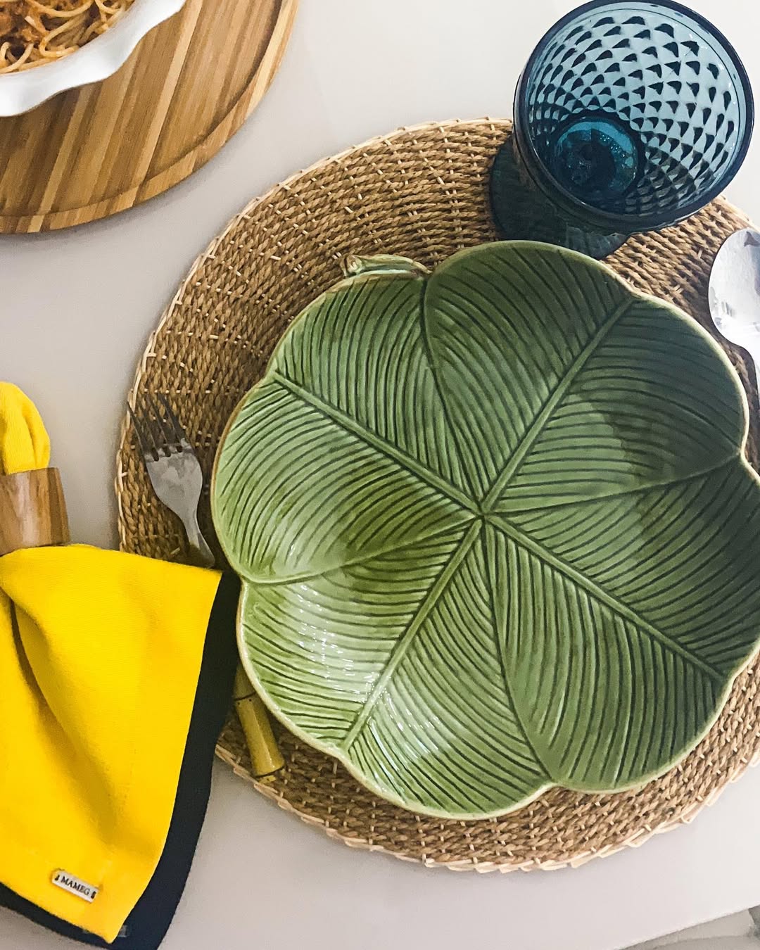 Mesa posta com prato verde em formato de folha sobre jogo americano de palha, copo azul texturizado, talheres e guardanapo amarelo com borda preta