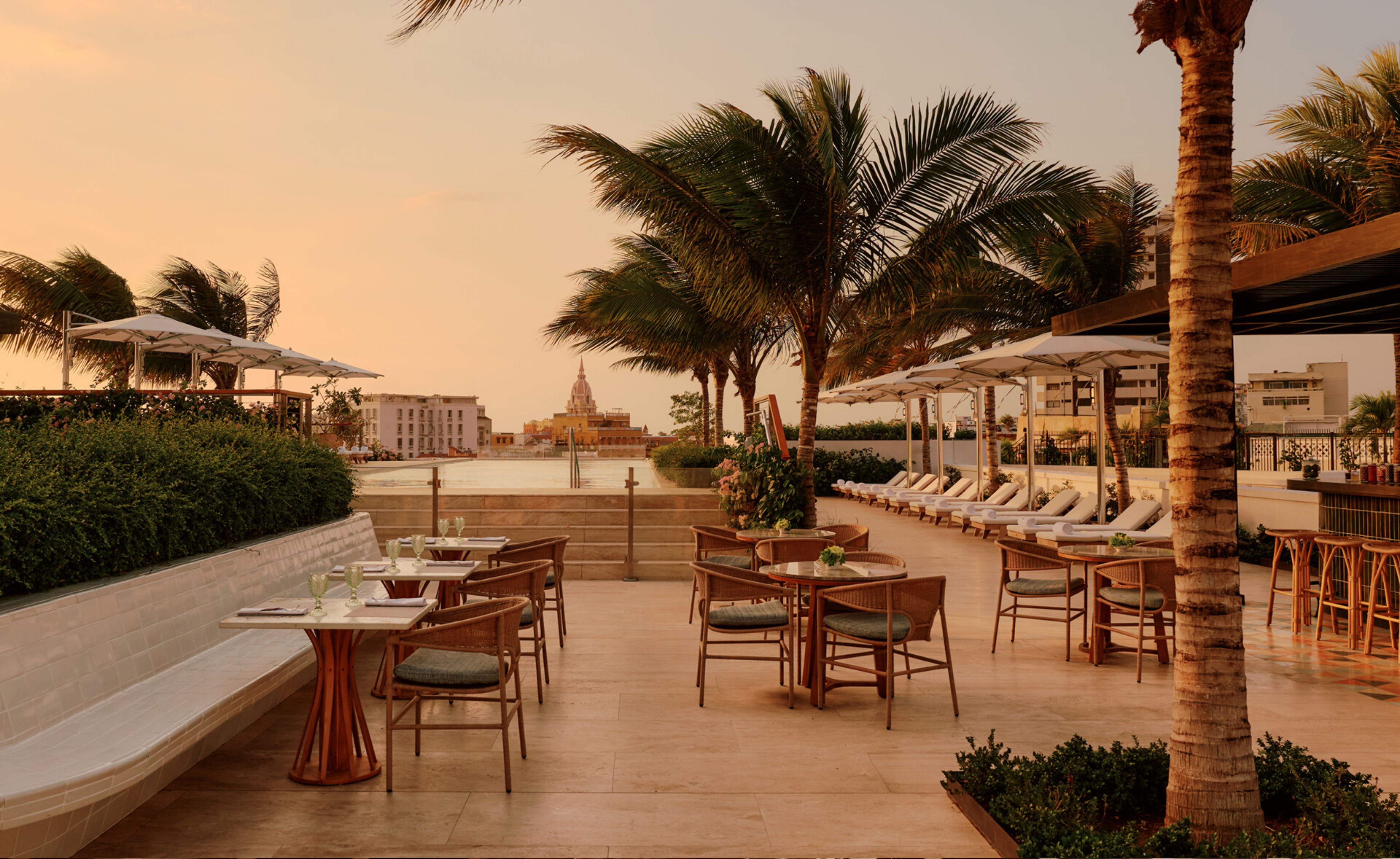 Terra&ccedil;o de um resort ao p&ocirc;r do sol, com mesas e cadeiras de vime, espregui&ccedil;adeiras brancas sob guarda-s&oacute;is, palmeiras e arbustos. Ao fundo, um corpo d&aacute;gua e edif&iacute;cios antigos de uma cidade costeira, incluindo uma c&uacute;pula. O c&eacute;u est&aacute; em tons de laranja e rosa