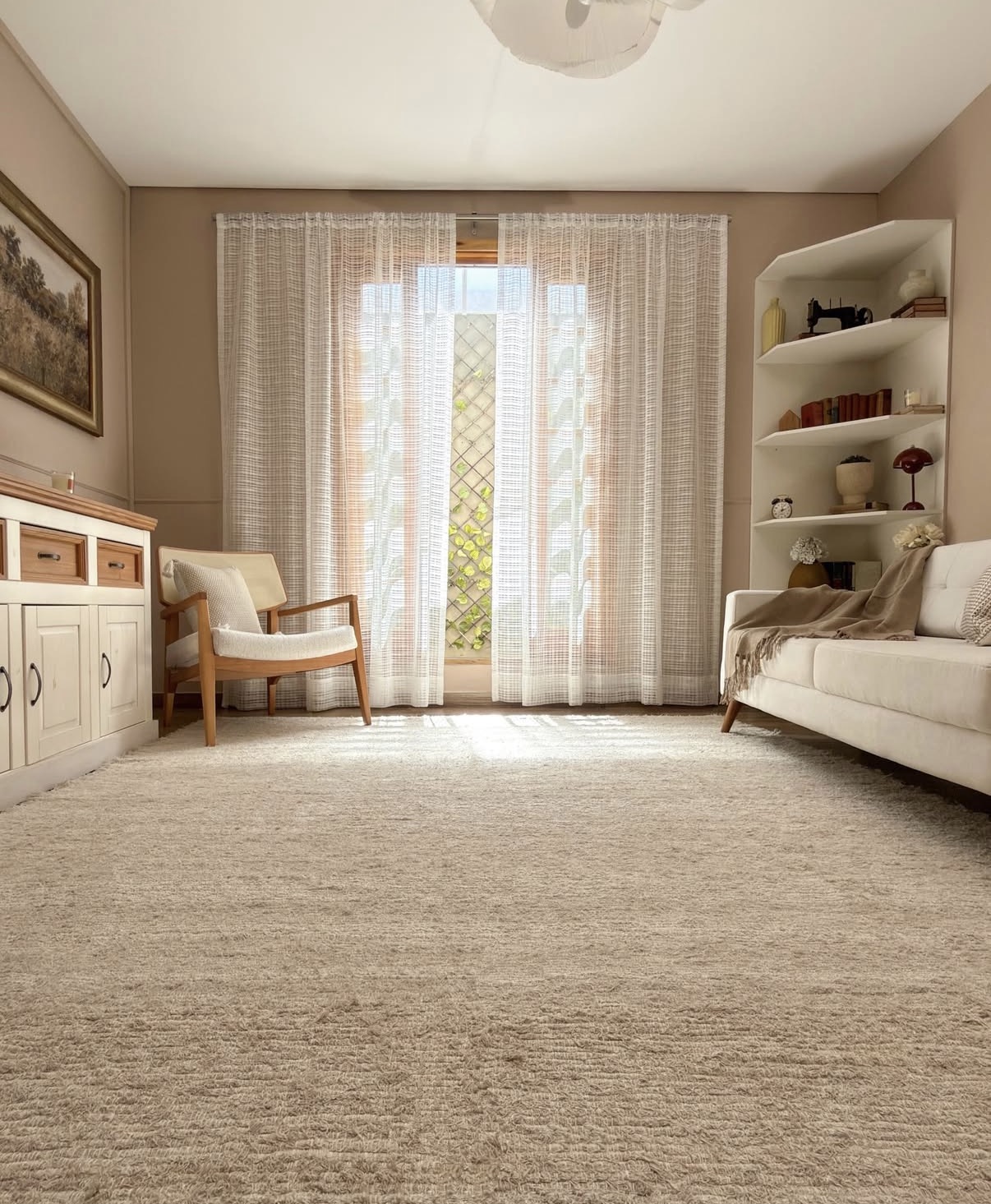Sala de estar clara com carpete bege, sof&aacute; branco com manta marrom, poltrona de madeira clara, estante de canto com livros e objetos decorativos, e janela com cortinas transl&uacute;cidas que revelam folhagens verdes