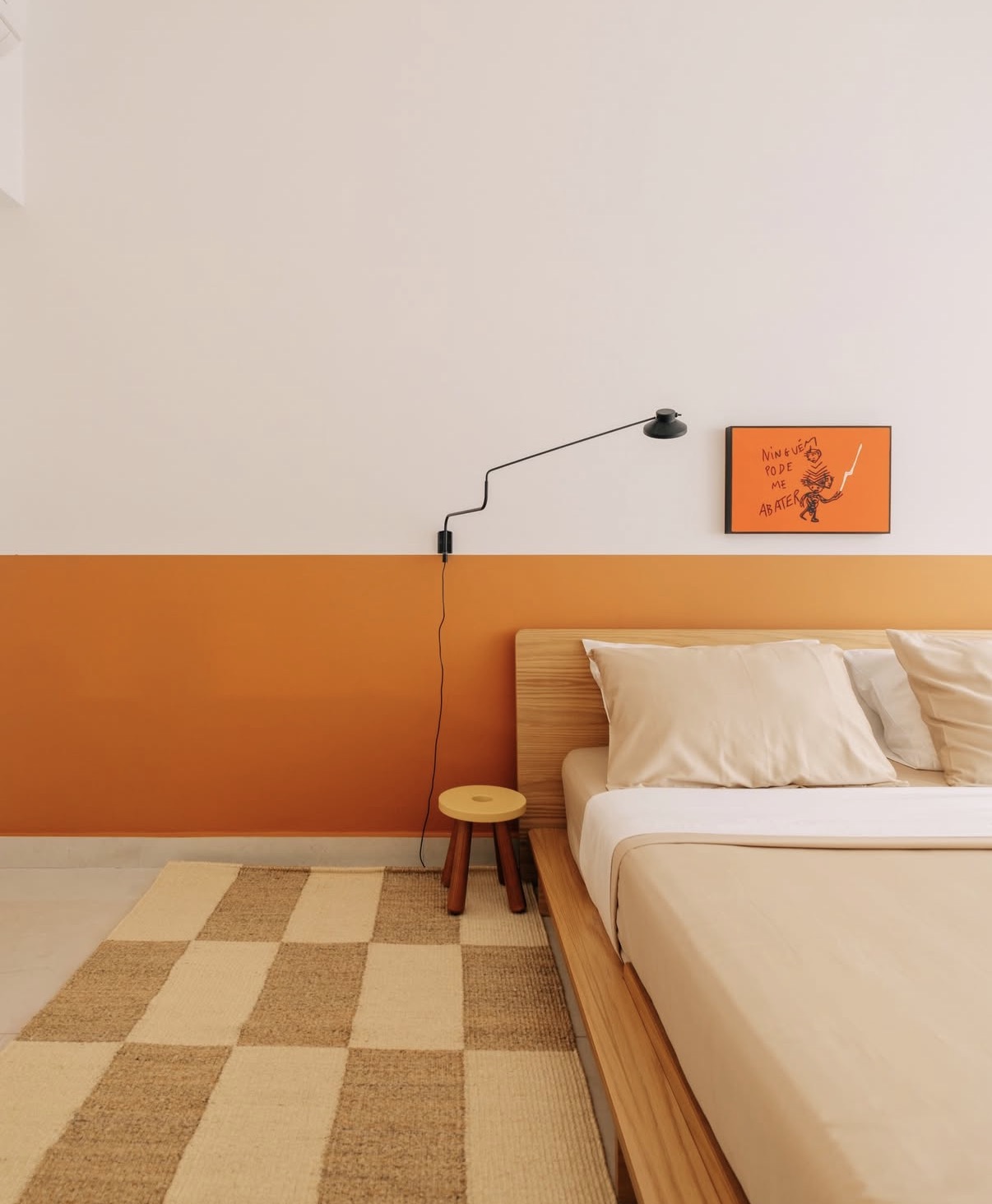 Quarto moderno com parede branca e faixa laranja, cama de madeira clara com len&ccedil;&oacute;is bege, banquinho de madeira e tapete xadrez bege e marrom. Na parede, um abajur preto e um quadro laranja com a frase Ningu&eacute;m pode me abater e um desenho de um guerreiro