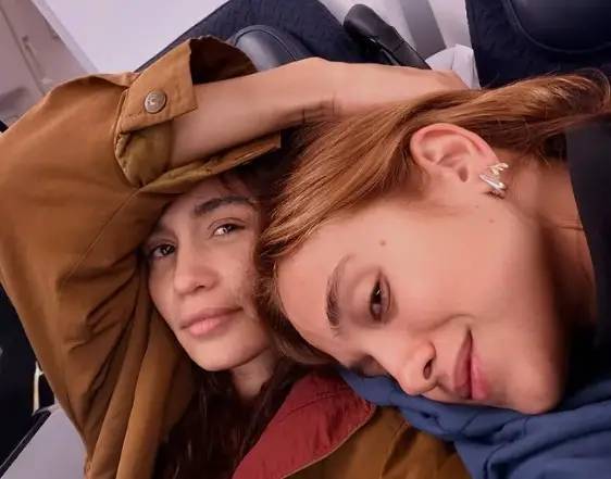 Duas mulheres jovens, uma com cabelos castanhos cacheados e outra com cabelos ruivos lisos, deitadas juntas em um avi&atilde;o. A mulher de cabelo castanho usa uma jaqueta marrom e olha para a c&acirc;mera com uma express&atilde;o neutra. A mulher de cabelo ruivo, com um brinco prateado em formato de asa, sorri suavemente enquanto apoia a cabe&ccedil;a no ombro da outra, vestindo uma blusa azul. Ambas parecem relaxadas e confort&aacute;veis