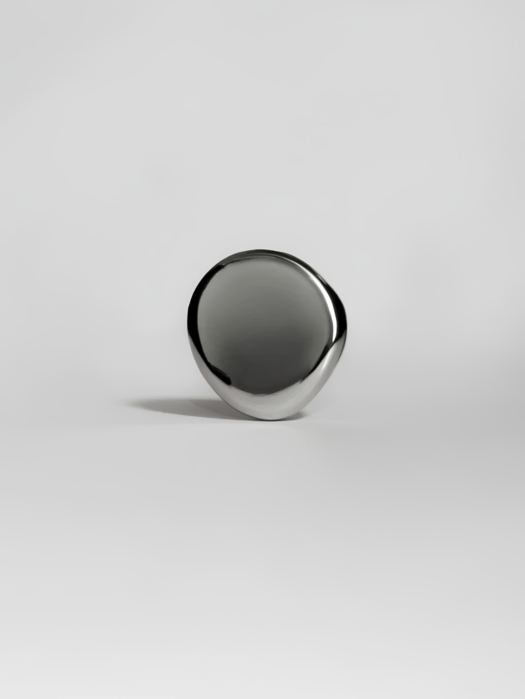 Objeto met&aacute;lico prateado, liso e brilhante, com formato oval irregular, apoiado em uma superf&iacute;cie branca. Uma sombra escura e alongada se projeta &agrave; esquerda, criando contraste e profundidade. A ilumina&ccedil;&atilde;o destaca o reflexo na superf&iacute;cie do objeto.