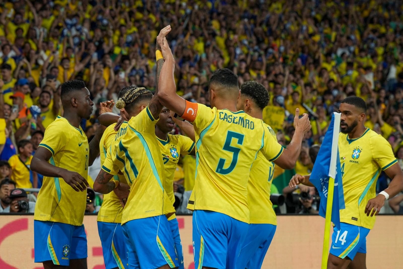 Jogadores da sele&ccedil;&atilde;o brasileira de futebol, com camisas amarelas e shorts azuis, comemoram um gol em campo, abra&ccedil;ados e com os bra&ccedil;os levantados. A torcida, vestida de amarelo, vibra ao fundo