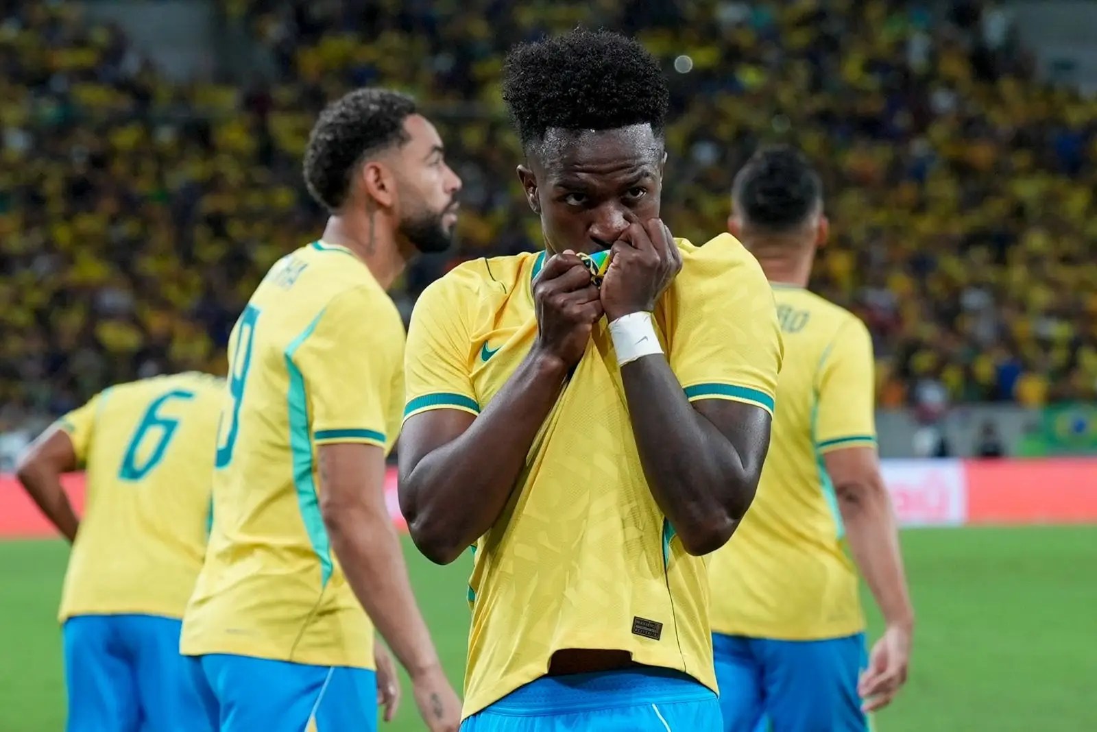 Jogador de futebol Vinicius Jr, com cabelo crespo, vestindo camisa amarela da seleção brasileira, com as mãos cobrindo a boca, em campo, com outros jogadores e torcida ao fundo