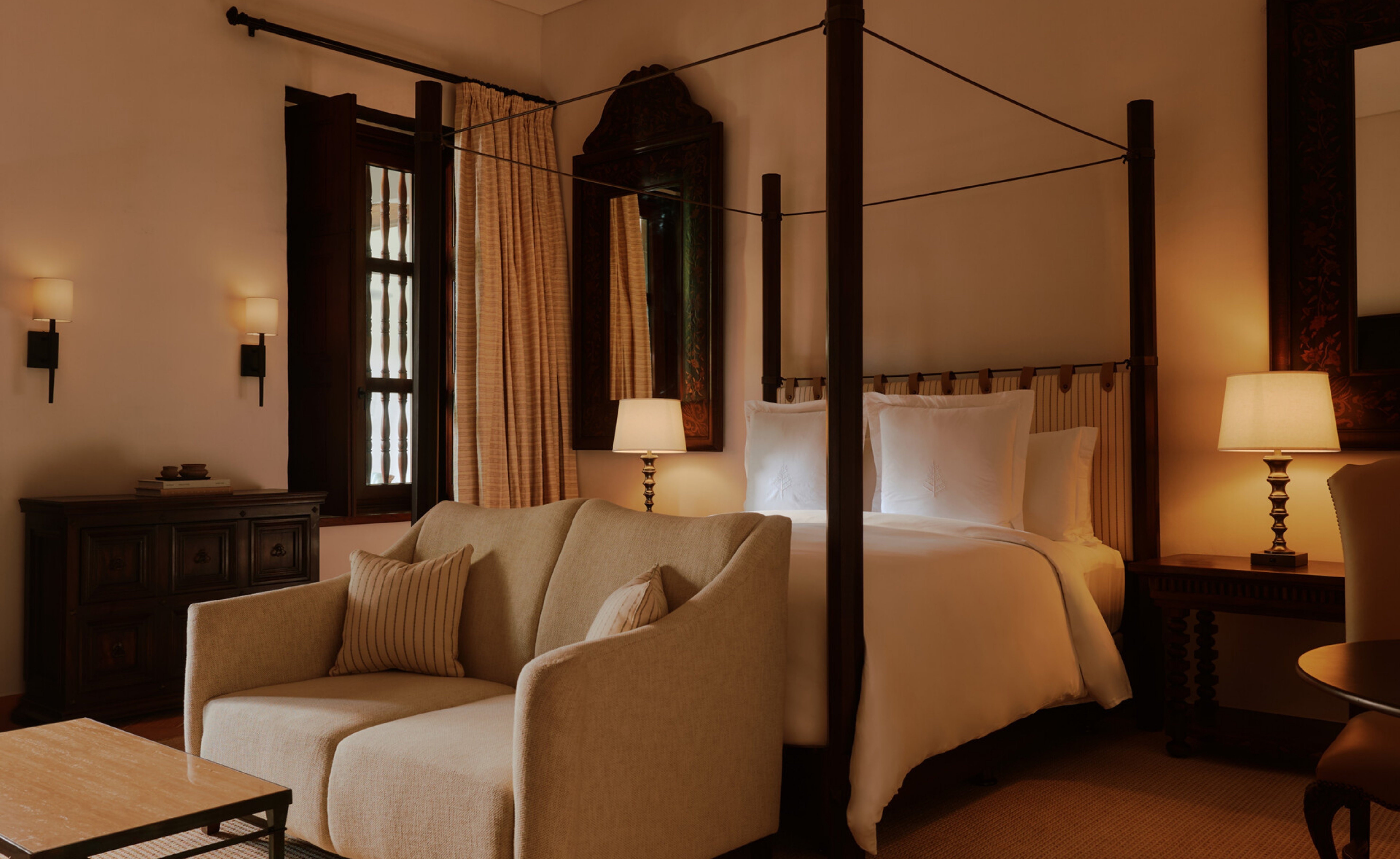 Quarto de hotel luxuoso com cama de dossel, len&ccedil;&oacute;is brancos, sof&aacute; bege com almofadas listradas, mesa de centro de madeira clara e abajures acesos. Uma janela escura com persianas e cortinas claras aparece ao fundo, e um espelho grande com moldura escura reflete o ambiente. O quarto tem ilumina&ccedil;&atilde;o quente e aconchegante