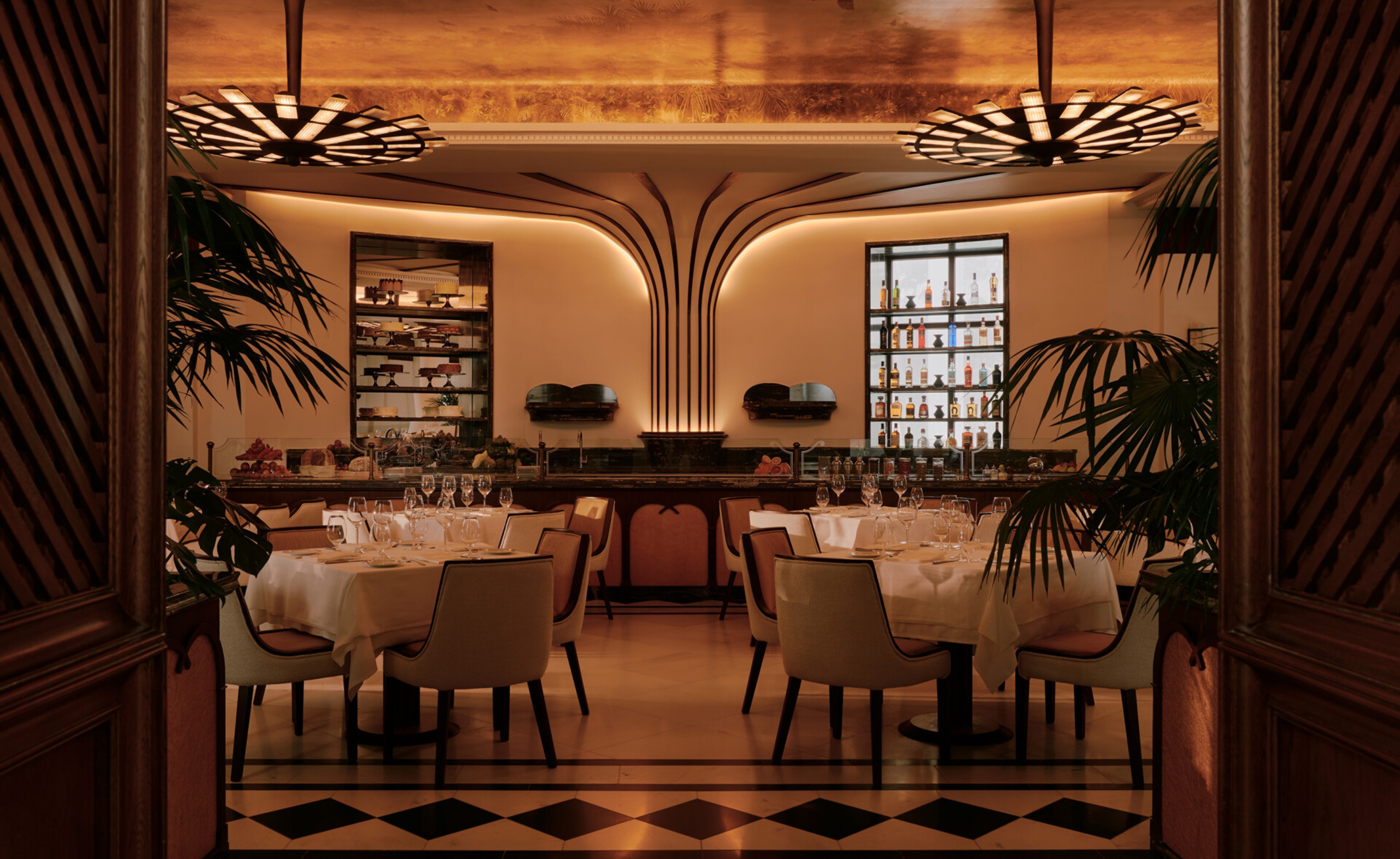 Interior de um restaurante luxuoso com mesas postas, cadeiras estofadas claras e piso xadrez. Ao fundo, um bar com garrafas de bebidas e prateleiras com doces. Plantas grandes decoram o ambiente, iluminado por lustres art&iacute;sticos e luzes indiretas, criando uma atmosfera sofisticada