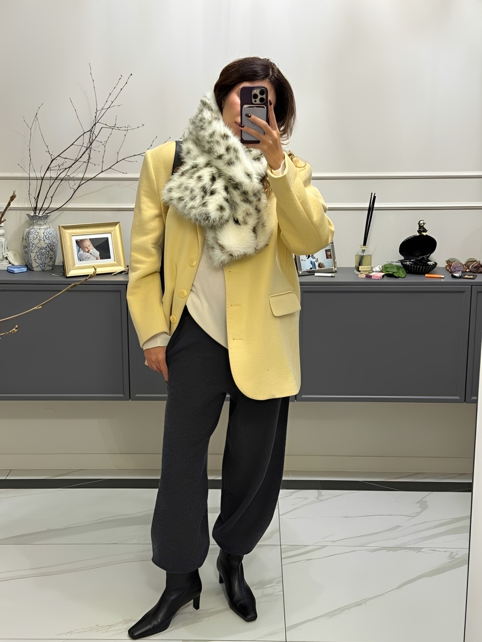 Pessoa de p&eacute;, tirando selfie no espelho, usando blazer amarelo, cal&ccedil;a escura e cachecol de pele sint&eacute;tica com estampa de leopardo. Ao fundo, um aparador cinza com objetos decorativos e um vaso com galhos secos