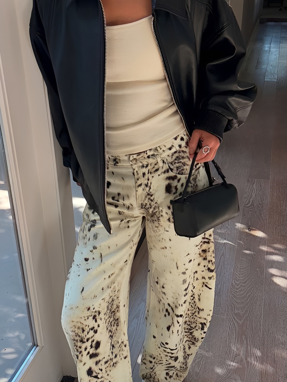 Pessoa com jaqueta preta de couro, regata creme e cal&ccedil;a larga com estampa de manchas marrons e pretas, segurando uma bolsa pequena preta. O cen&aacute;rio &eacute; um ambiente interno com piso de madeira e uma porta branca ao lado esquerdo, com luz solar projetando sombras no ch&atilde;o