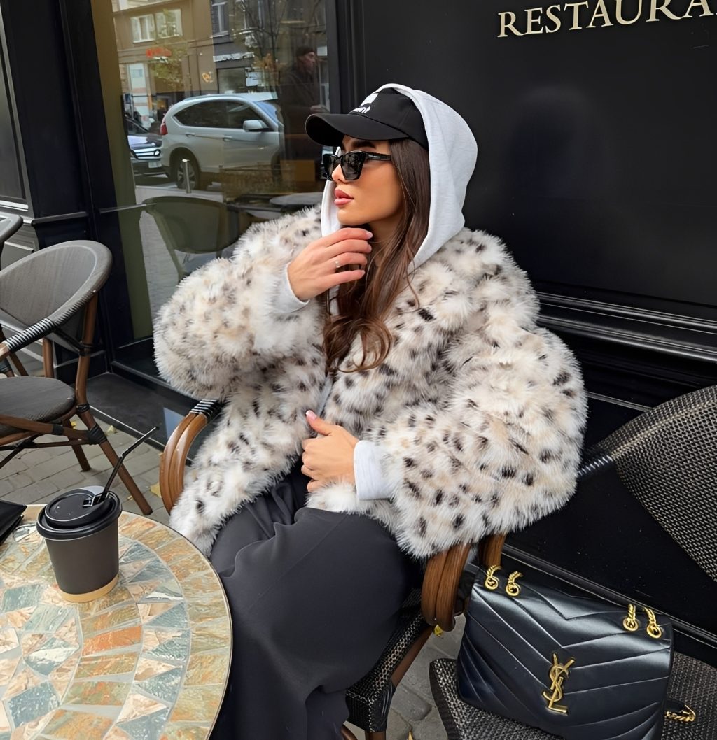 Mulher jovem sentada em uma cadeira de caf&eacute; ao ar livre, usando casaco de pele sint&eacute;tica com estampa de leopardo, moletom branco com capuz, bon&eacute; preto e &oacute;culos escuros. Ela olha para a esquerda, com uma bolsa preta ao lado e um copo de caf&eacute; na mesa de mosaico.
