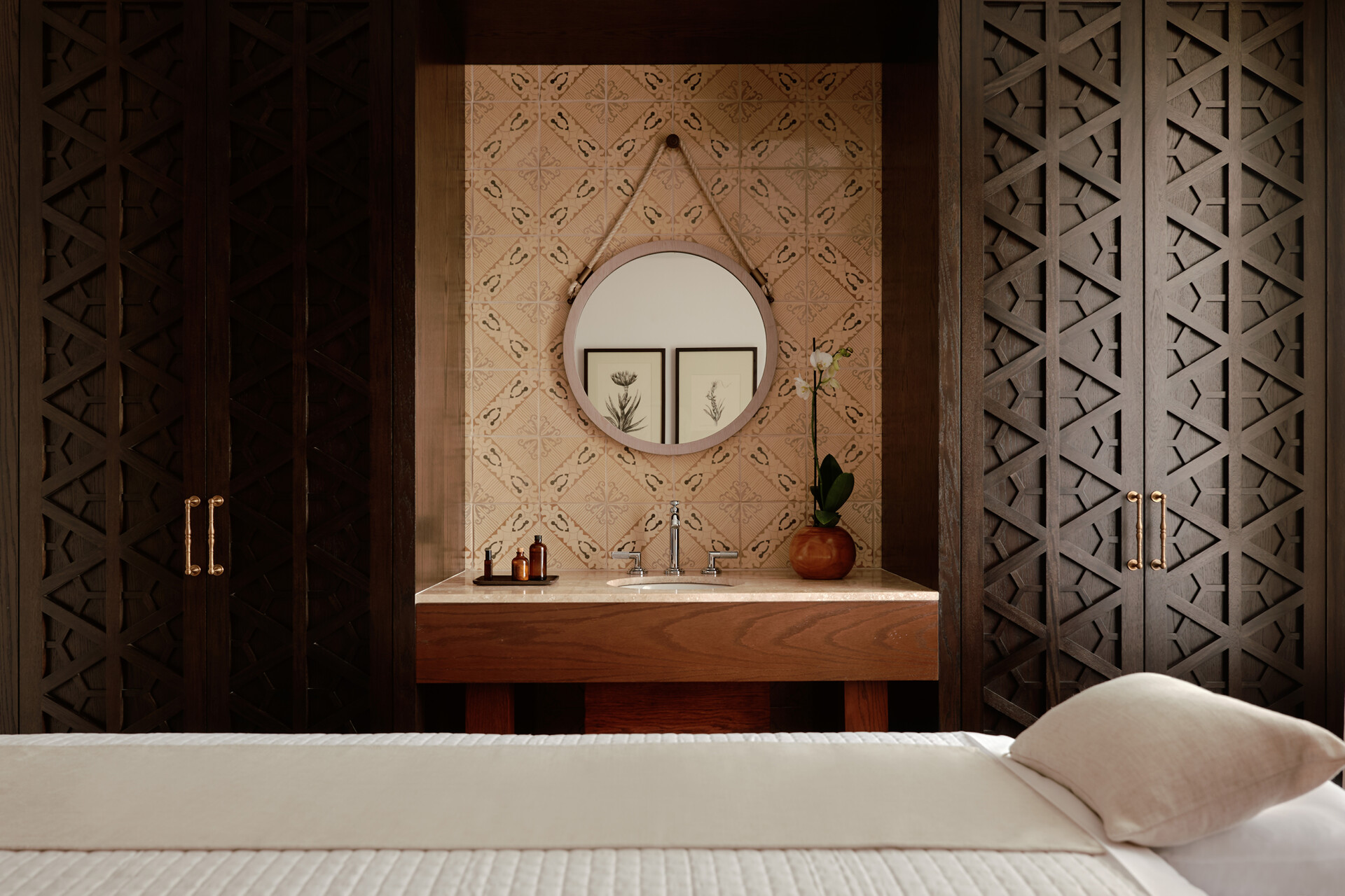 Um quarto de hotel com cama branca em primeiro plano, e ao fundo, um balc&atilde;o de madeira com pia, espelho redondo e azulejos bege. Arm&aacute;rios escuros com detalhes geom&eacute;tricos ladeiam o balc&atilde;o