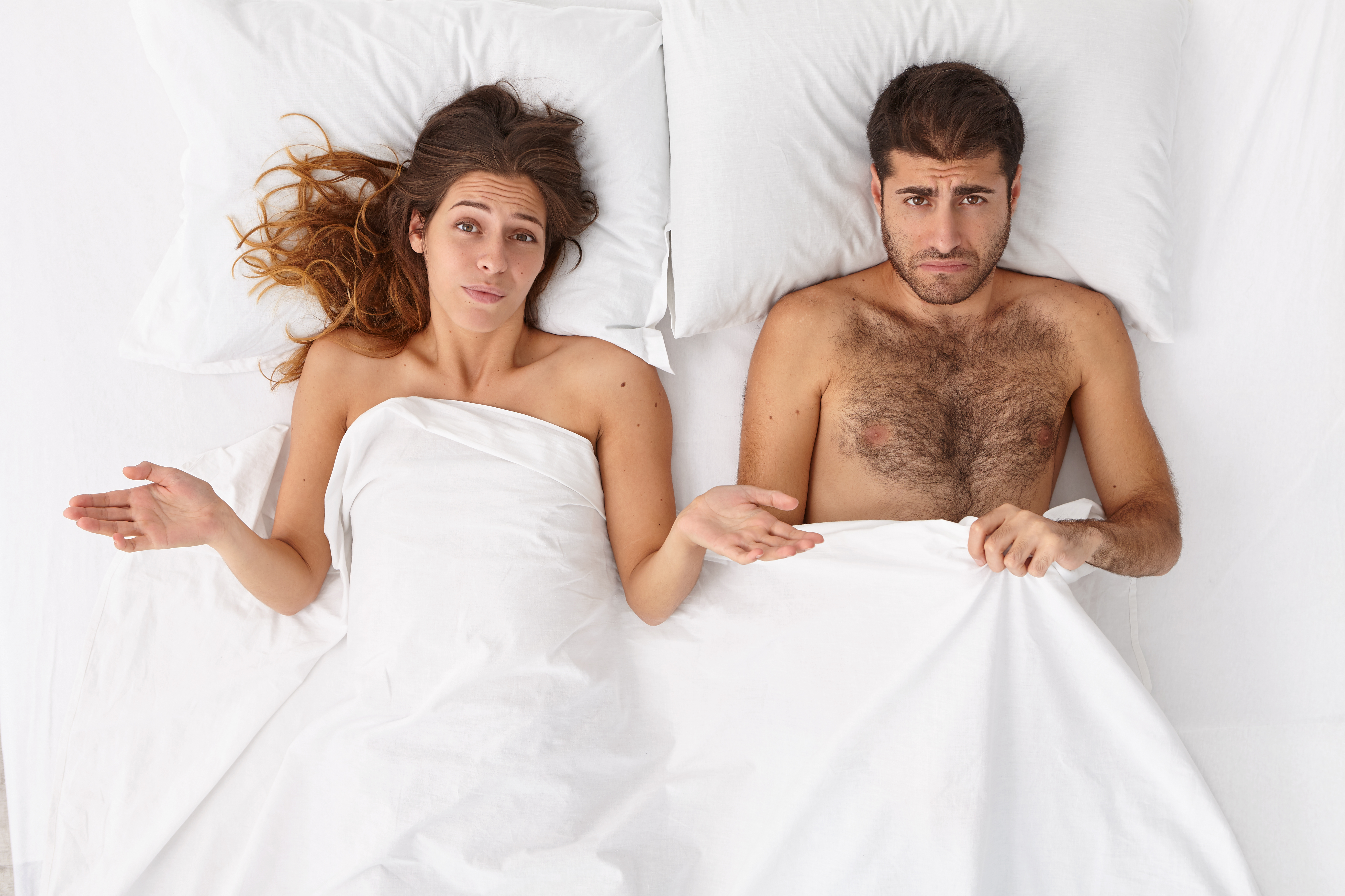 Casal deitado na cama com len&ccedil;&oacute;is brancos. A mulher, &agrave; esquerda, tem express&atilde;o confusa e m&atilde;os abertas. O homem, &agrave; direita, com peito peludo, parece chateado e segura o len&ccedil;ol. Ambos olham para cima