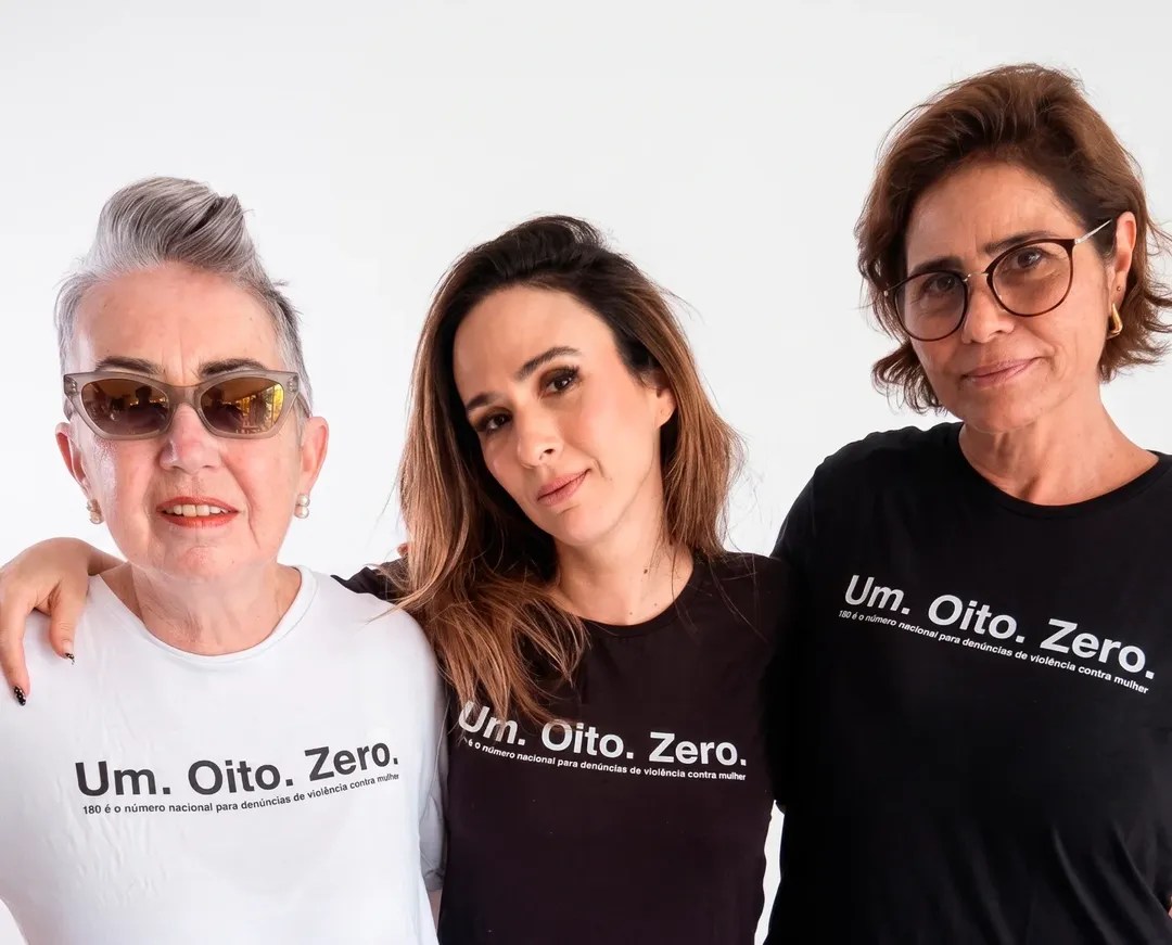 Três mulheres posam sorrindo, a da esquerda de óculos escuros e cabelo grisalho, a do centro com cabelo castanho e a da direita de óculos de grau e cabelo castanho. Todas vestem camisetas com a frase Um. Oito. Zero. 180 é o número nacional para denúncias de violência contra mulher