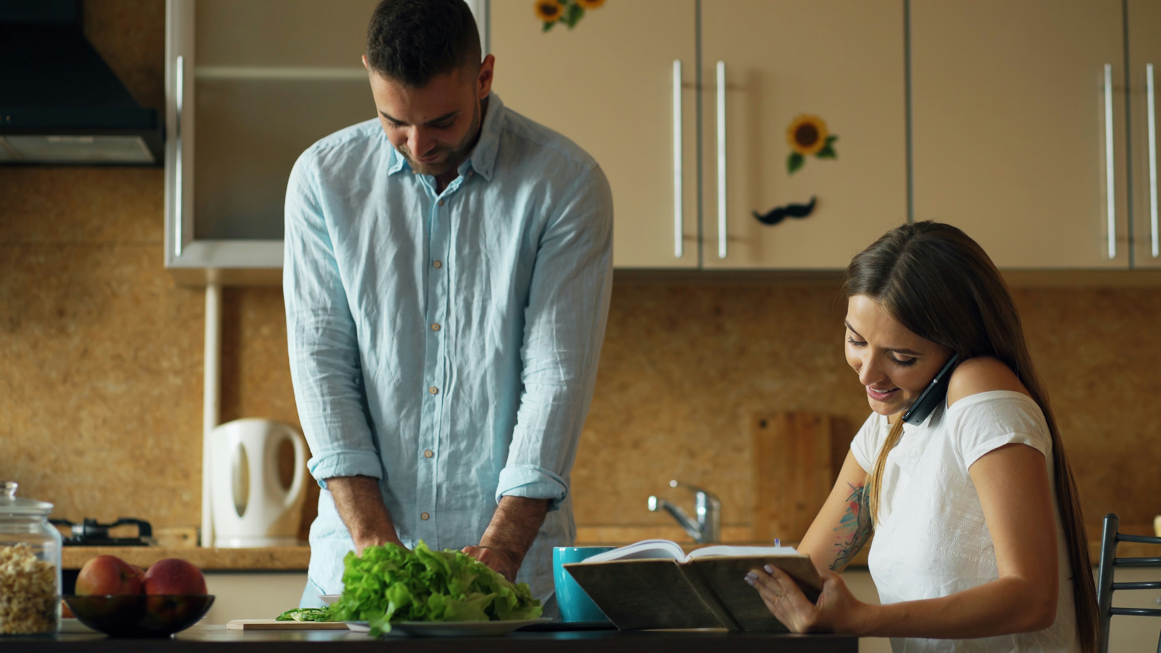 Homem de camisa azul picando alface na cozinha, enquanto mulher de blusa branca sorri ao telefone, lendo um livro de receitas
