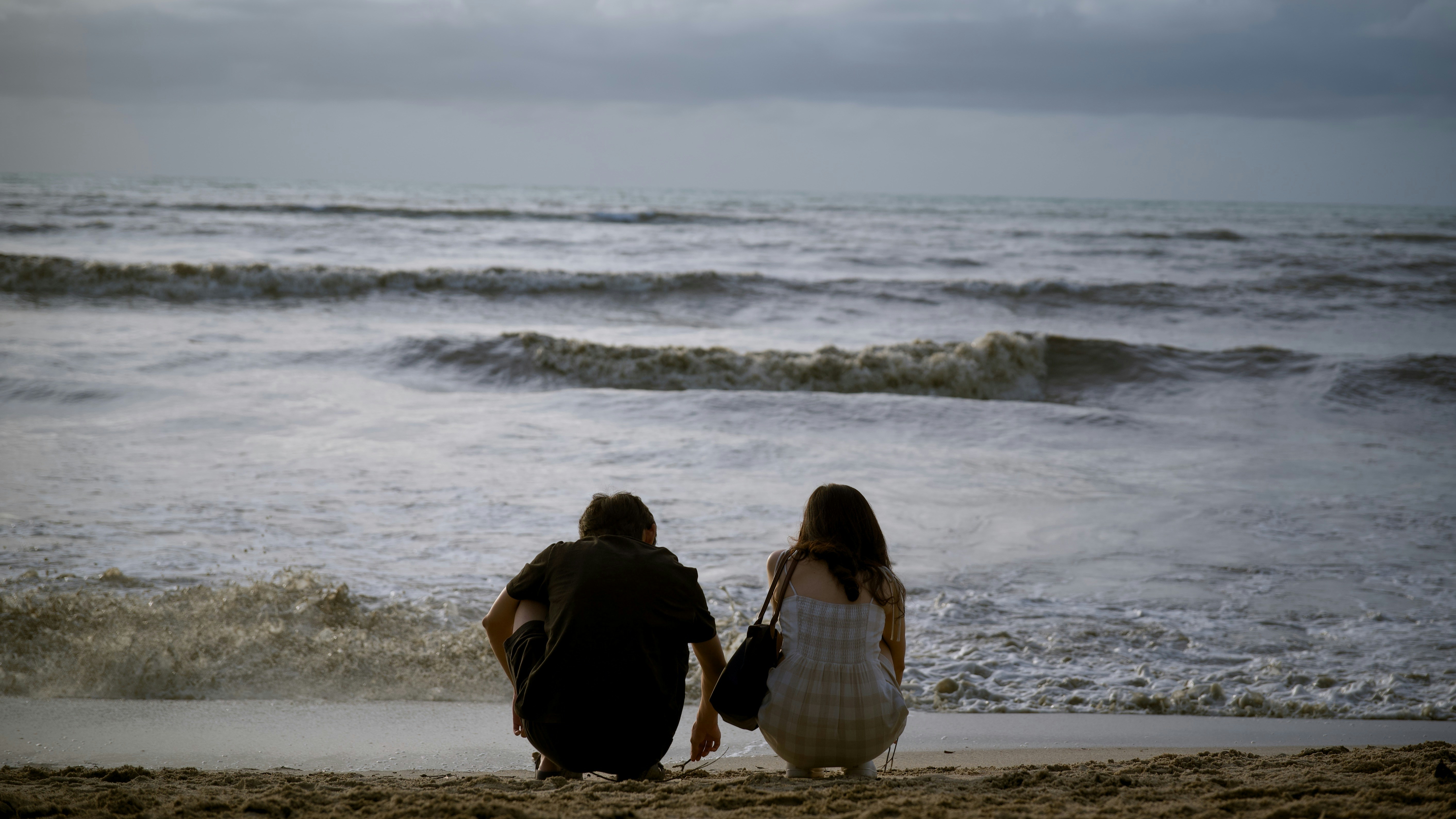 Um casal agachado na areia da praia, de costas, observando as ondas do mar sob um c&eacute;u nublado. O homem veste camiseta escura e a mulher um vestido claro, com uma bolsa preta no ombro.
