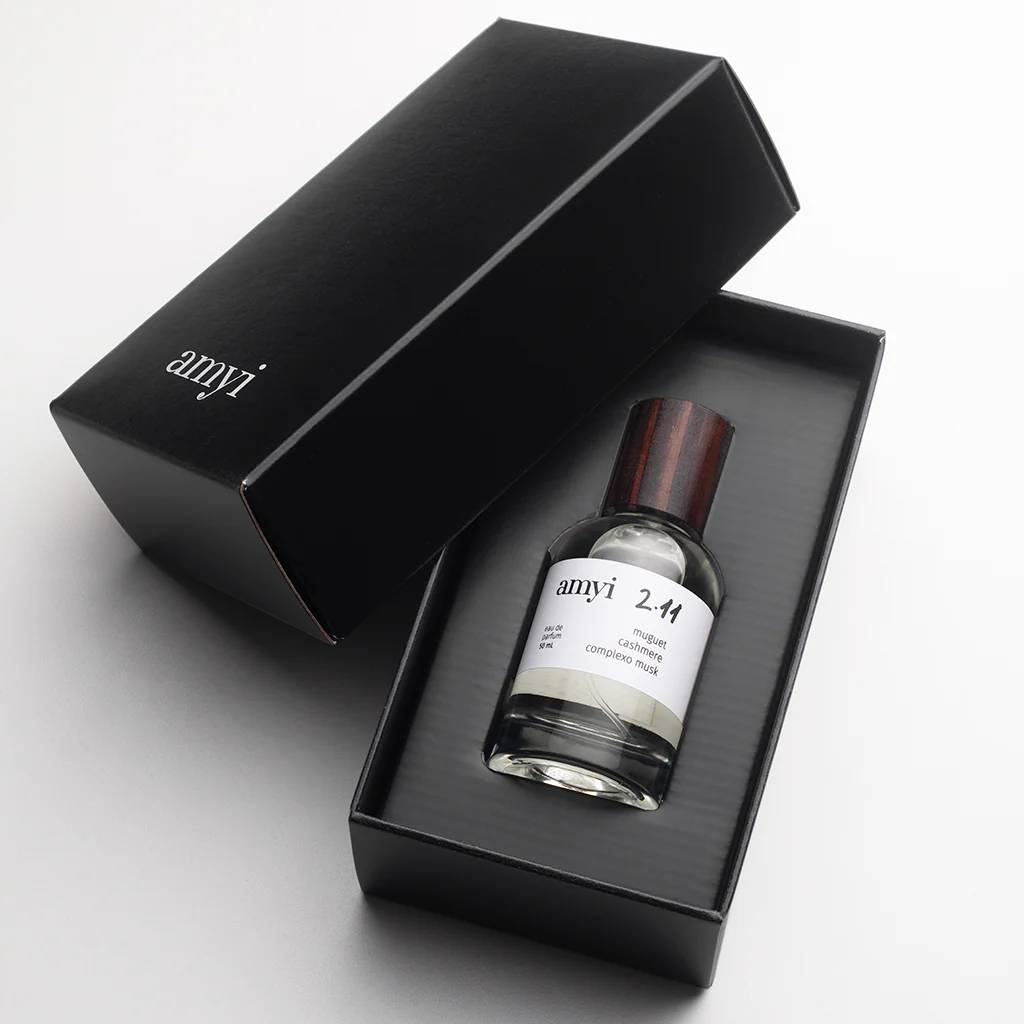 Frasco de perfume transparente com tampa de madeira escura dentro de uma caixa preta aberta, com o logo amyi em branco na tampa e no r&oacute;tulo do frasco, que tamb&eacute;m indica 2.11 muguet cashmere complexo musk e eau de parfum 50 mL