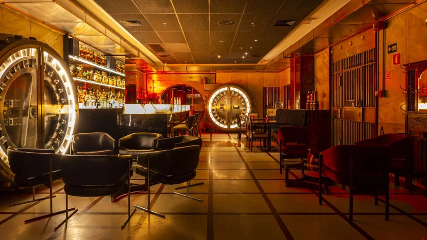 Interior de um bar com ilumina&ccedil;&atilde;o &acirc;mbar, decorado como um cofre de banco antigo. H&aacute; um balc&atilde;o com garrafas de bebidas, poltronas de couro pretas e sof&aacute;s de veludo vermelho. Duas grandes portas de cofre circulares, uma delas com um rel&oacute;gio, dominam o ambiente, e as paredes s&atilde;o revestidas com grades met&aacute;licas e pain&eacute;is dourados. O ch&atilde;o tem um padr&atilde;o de quadrados com linhas escuras.