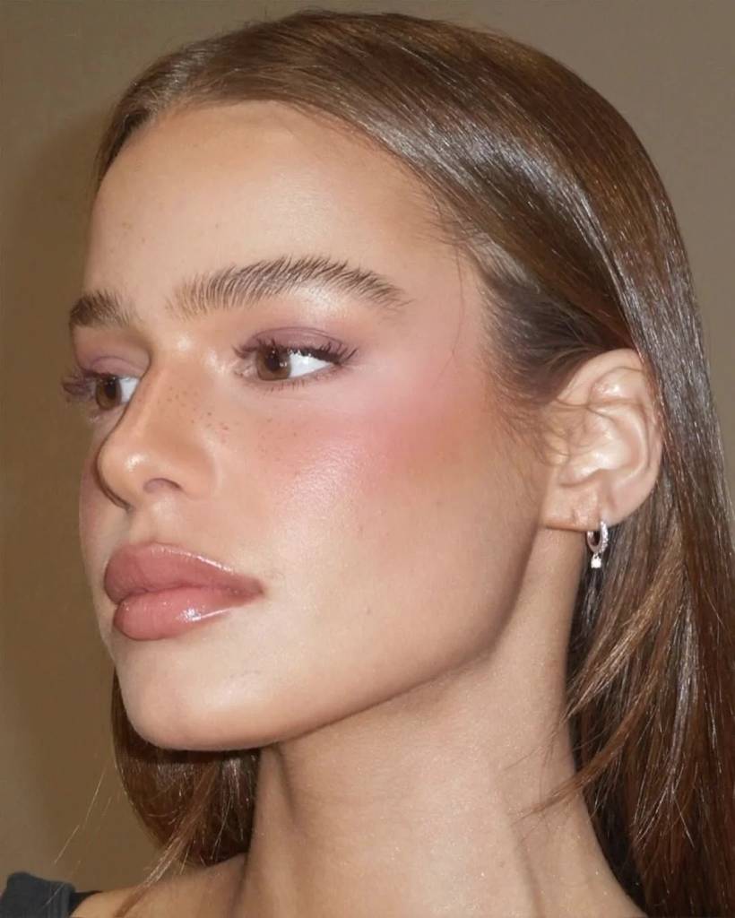 Rosto de mulher jovem com pele clara, sardas no nariz, maquiagem rosa nos olhos e bochechas, l&aacute;bios com gloss e cabelo castanho solto. Ela usa um brinco de argola prateado com um pequeno pingente quadrado