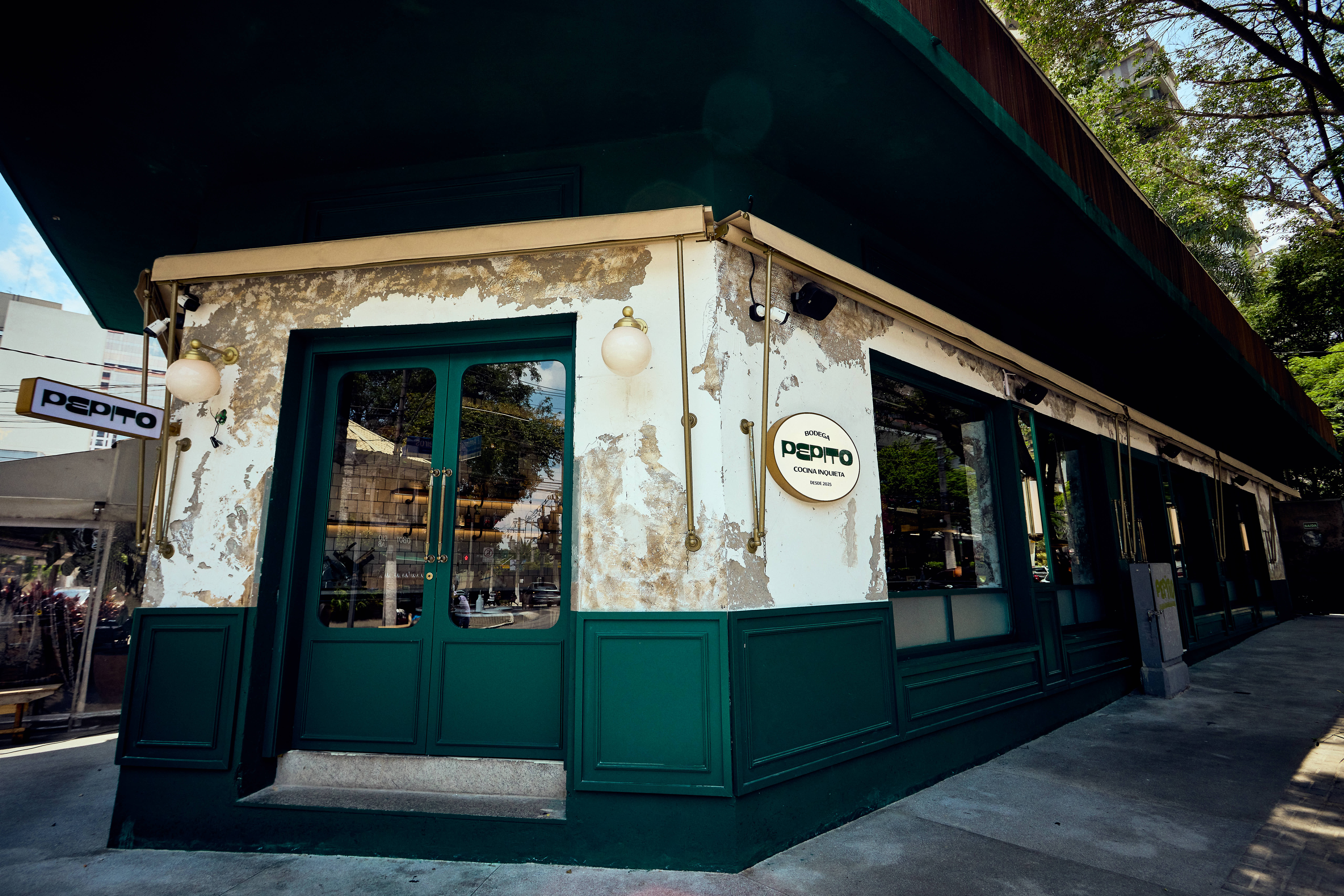 Fachada de um restaurante chamado Bodega Pepito Cozinha Indiscreta, com paredes brancas descascadas e detalhes em verde escuro, incluindo a porta e as molduras das janelas. H&aacute; um toldo verde escuro e &aacute;rvores ao fundo, sob um c&eacute;u azul