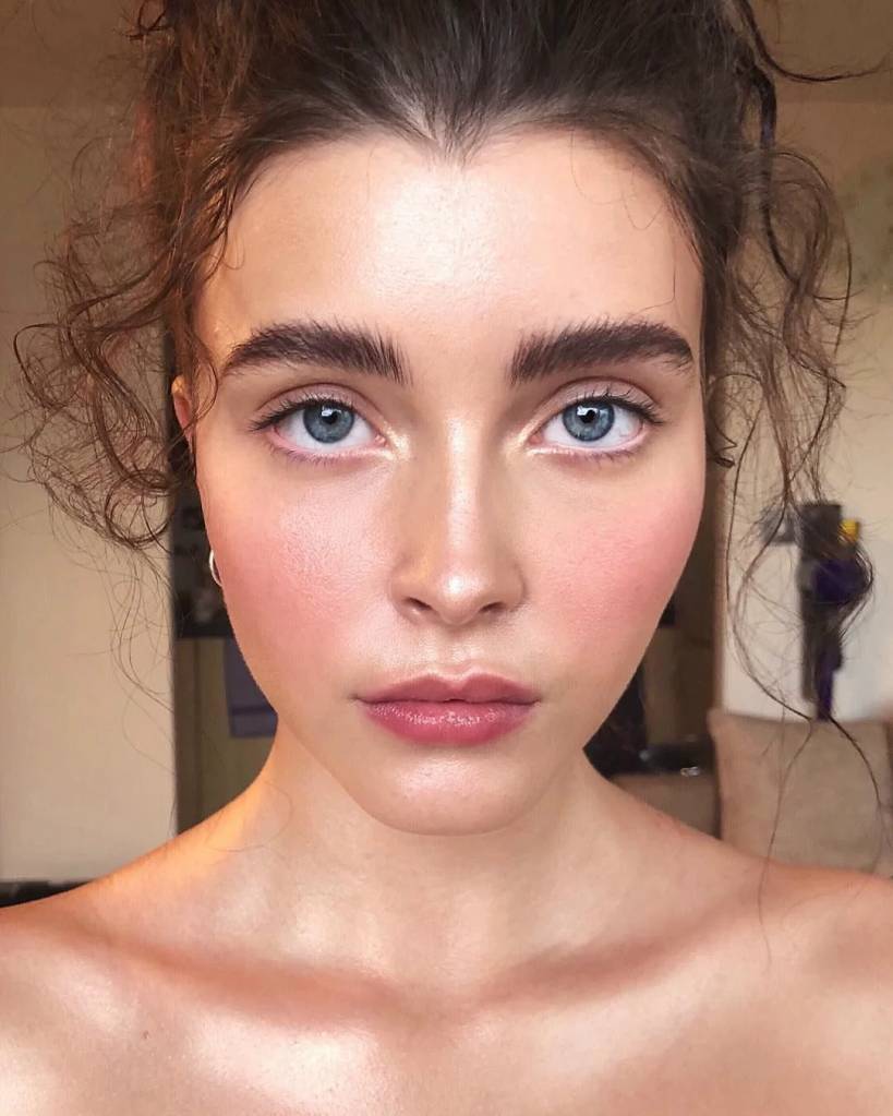Rosto de uma mulher jovem com pele clara, olhos azuis intensos e sobrancelhas grossas. Ela tem bochechas rosadas, l&aacute;bios com batom rosa e cabelo castanho-escuro preso, com alguns fios soltos. Seus ombros est&atilde;o expostos, sugerindo uma blusa tomara que caia ou sem al&ccedil;as. A express&atilde;o &eacute; neutra, olhando diretamente para frente