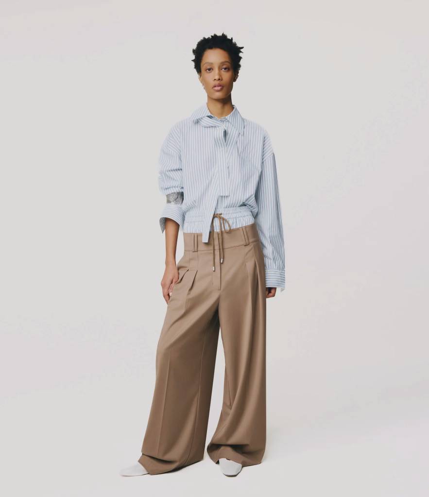 Mulher de pele escura e cabelo curto, vestindo camisa listrada azul e branca com amarra&ccedil;&atilde;o na cintura, cal&ccedil;a pantalona marrom e sapatos brancos, posando em fundo branco
