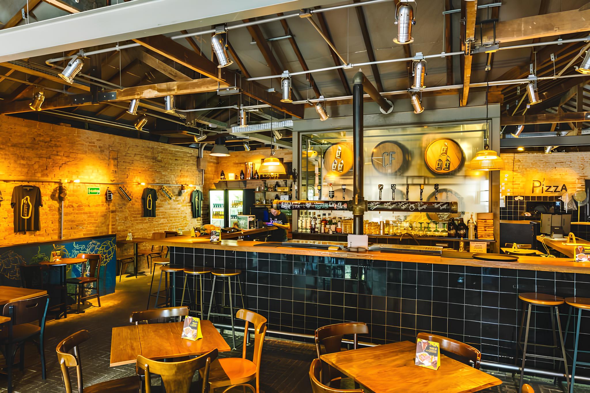 Interior de um bar com balc&atilde;o de azulejos pretos e bancos altos, mesas de madeira com cadeiras e paredes de tijolos &agrave; vista. No teto, vigas de madeira e ilumina&ccedil;&atilde;o industrial com spots. Ao fundo, um balc&atilde;o com torneiras de chope e garrafas, e um letreiro luminoso Pizza