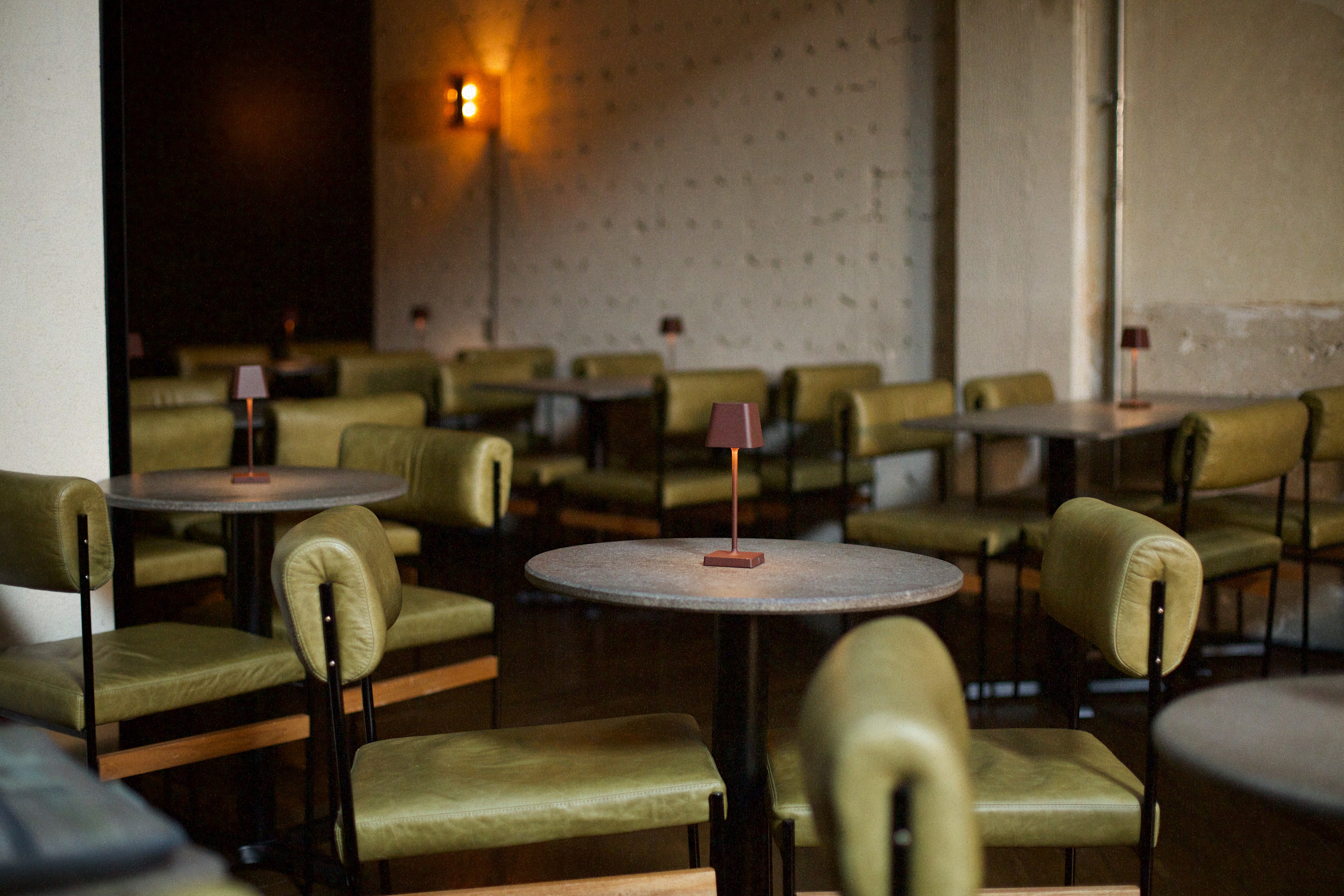 Interior de um restaurante ou bar com mesas redondas e cadeiras estofadas em couro verde-oliva. Pequenos abajures marrons iluminam cada mesa, e uma lumin&aacute;ria de parede laranja brilha ao fundo, criando uma atmosfera aconchegante