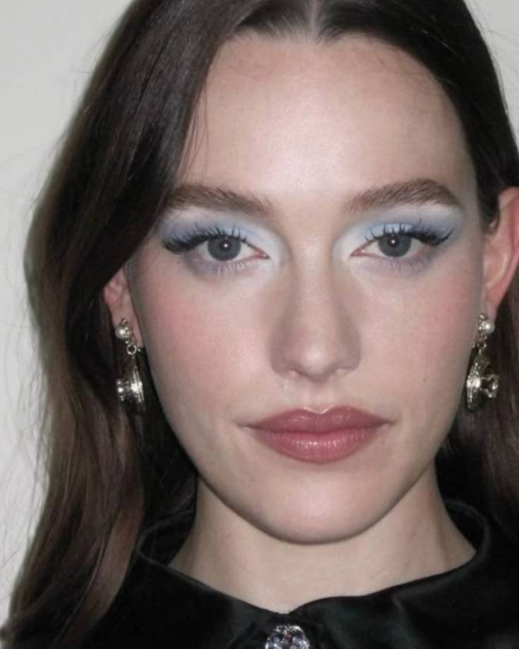 Rosto de mulher jovem com cabelo castanho escuro, maquiagem azul clara nos olhos e batom rosa. Ela usa brincos de p&eacute;rola e um colar com broche prateado