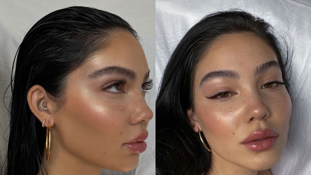 Mulher jovem com pele bronzeada, sardas, cabelo escuro molhado e maquiagem natural, com blush, iluminador e gloss labial. Usa brincos de argola dourados e piercing na cartilagem em formato de lua
