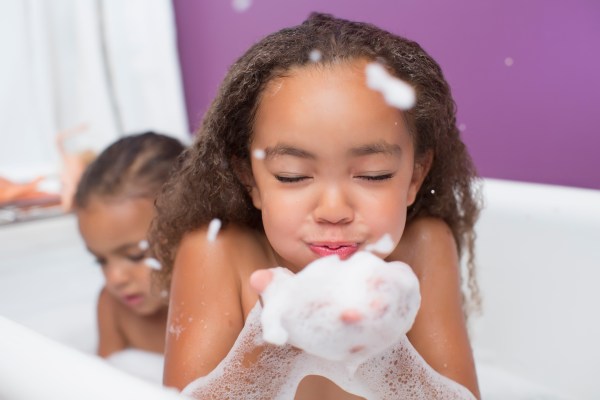 Menina de cabelos cacheados e pele morena com olhos fechados, soprando espuma das mãos na banheira. Outra criança aparece desfocada ao fundo, também na banheira, com o cabelo preso. O fundo é roxo e branco