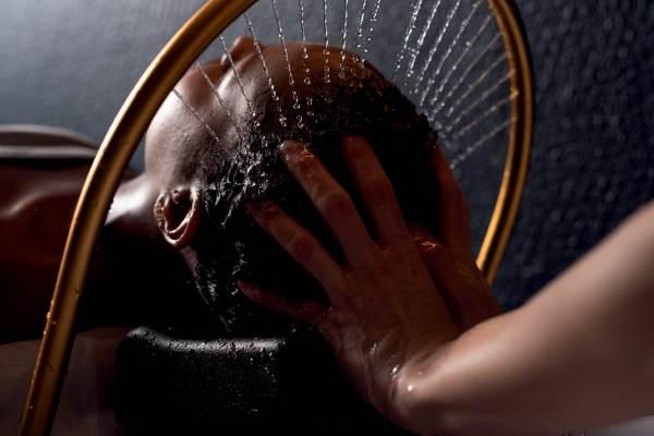 Pessoa de pele escura deitada de costas, recebendo um jato de água na cabeça de um chuveiro dourado curvo, enquanto uma mão clara massageia seu couro cabeludo. Gotas de água escorrem pelos cabelos e rosto, em um ambiente escuro