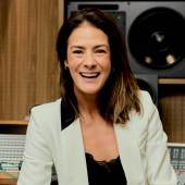 Mulher de cabelos castanhos, vestindo blazer branco e blusa preta de renda, sorri amplamente para a câmera em um estúdio de gravação com equipamentos de áudio e painéis de madeira ao fundo