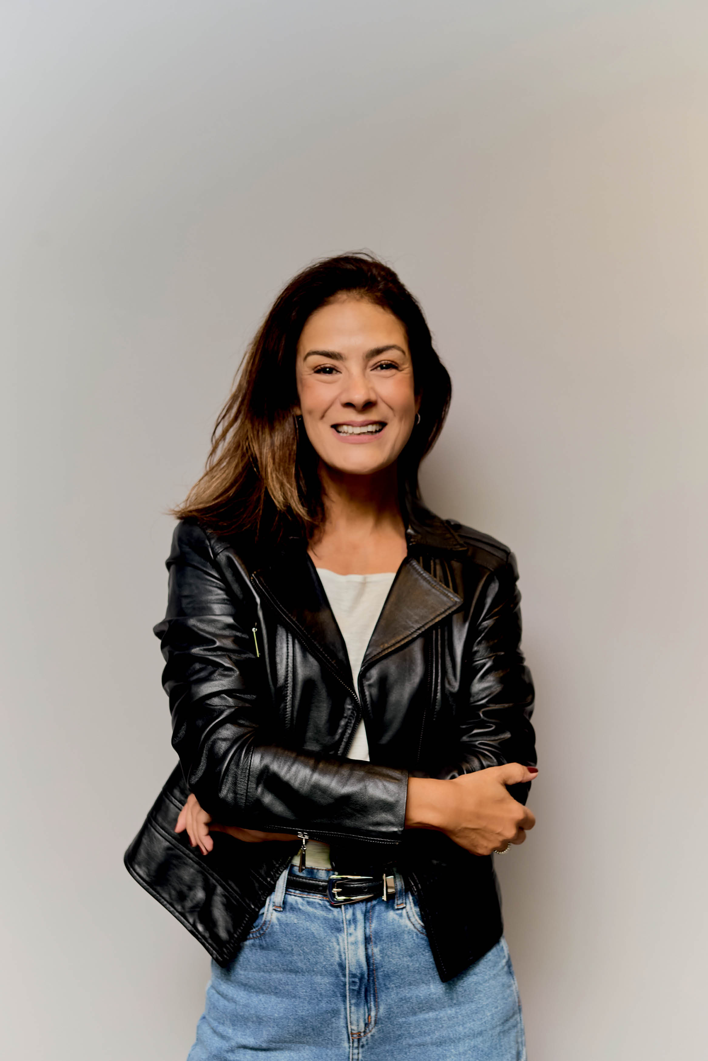 Mulher de cabelos castanhos, sorrindo, vestindo jaqueta de couro preta, camiseta branca e cal&ccedil;a jeans, bra&ccedil;os cruzados, em fundo cinza claro