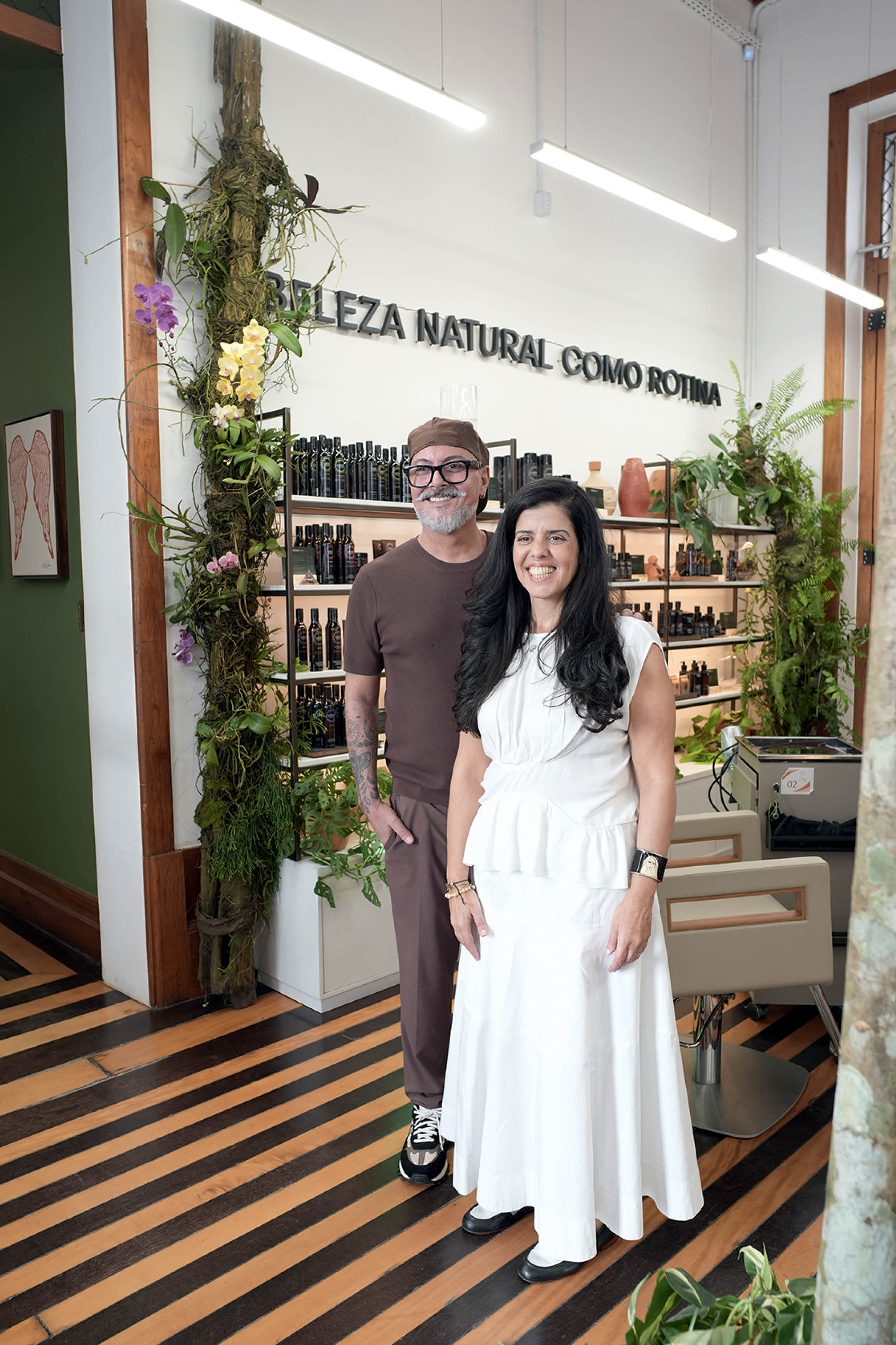 Um homem e uma mulher sorrindo, em p&eacute;, dentro de um estabelecimento com prateleiras de produtos e plantas. O homem usa bon&eacute; e camiseta marrons, a mulher veste um vestido branco. Ao fundo, na parede branca, l&ecirc;-se BELEZA NATURAL COMO ROTINA