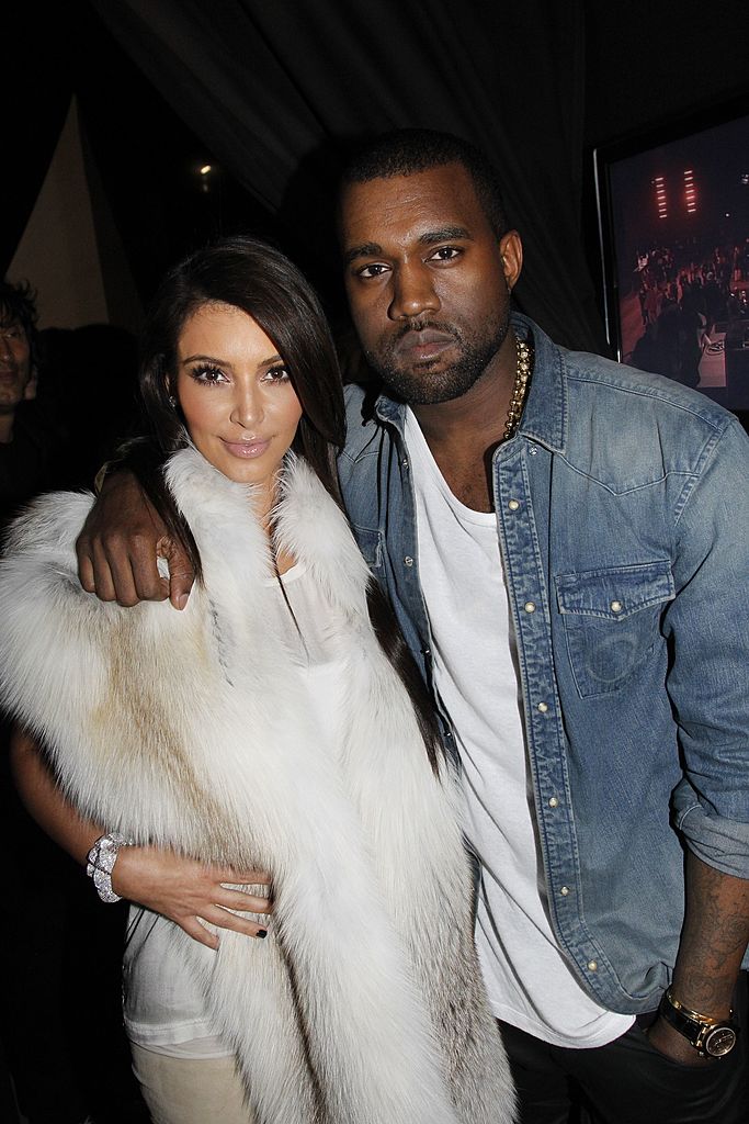 Kim Kardashian e Kanye West posam juntos em evento noturno. Kim sorri levemente, usando um casaco de pele branco e um bracelete brilhante. Kanye, com express&atilde;o s&eacute;ria, veste camisa jeans, camiseta branca e colar dourado. Ele a abra&ccedil;a pelo ombro.
