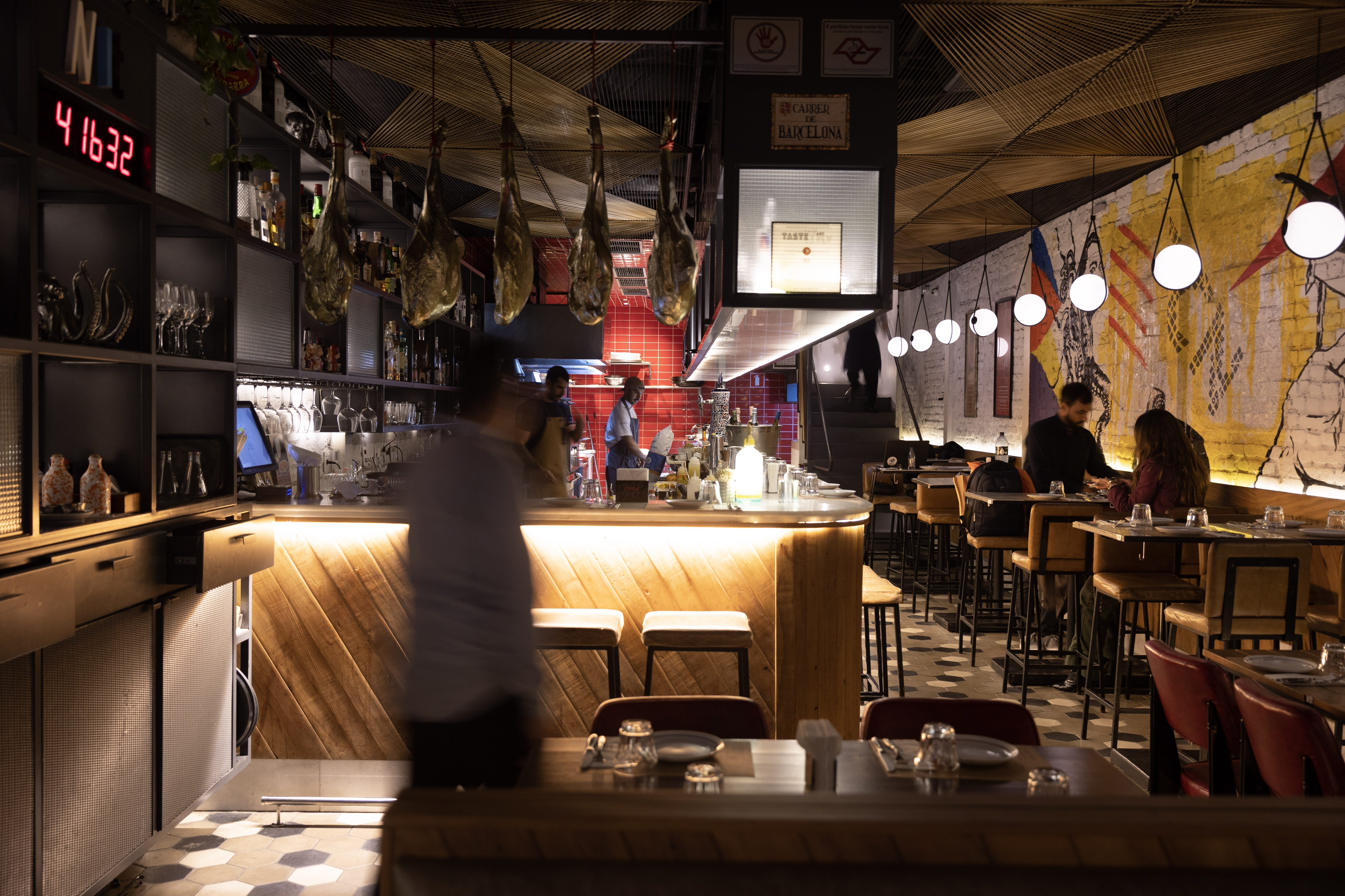 Interior de restaurante com ilumina&ccedil;&atilde;o baixa e ambiente moderno. Um balc&atilde;o de madeira iluminado &agrave; esquerda, com garrafas e ta&ccedil;as. Presuntos pendurados no teto. Ao fundo, uma cozinha com azulejos vermelhos e funcion&aacute;rios. &Agrave; direita, clientes sentados em mesas, com uma parede grafitada e lumin&aacute;rias esf&eacute;ricas penduradas