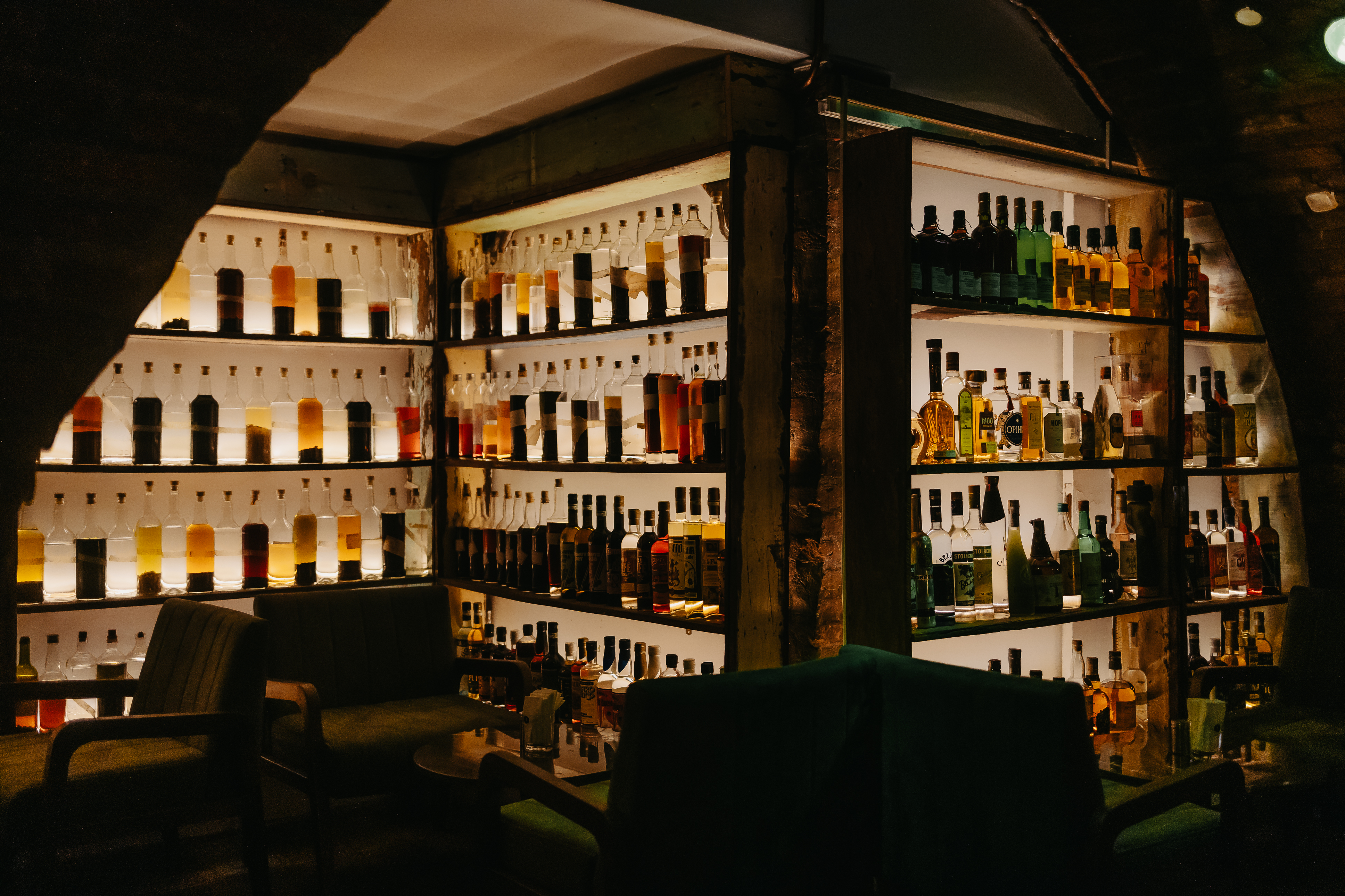 Interior de um bar escuro com prateleiras iluminadas exibindo centenas de garrafas de bebidas alco&oacute;licas de v&aacute;rias cores e tamanhos, e poltronas verdes escuras na frente