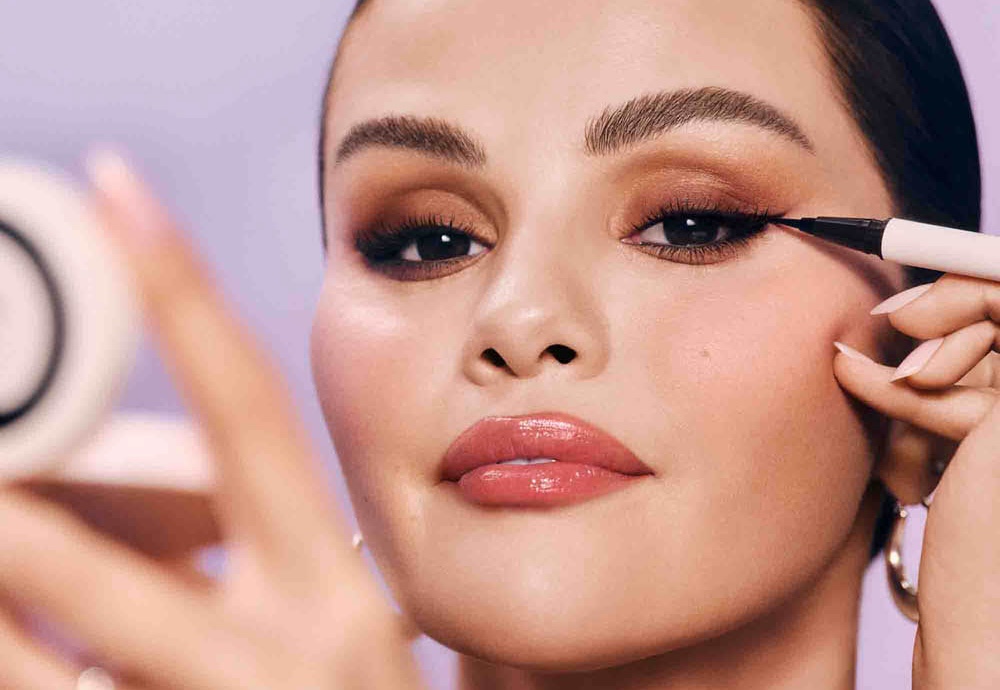 Selena Gomez aplicando delineador preto na pálpebra superior do olho direito, com maquiagem marrom nos olhos e batom rosa brilhante, contra fundo lilás
