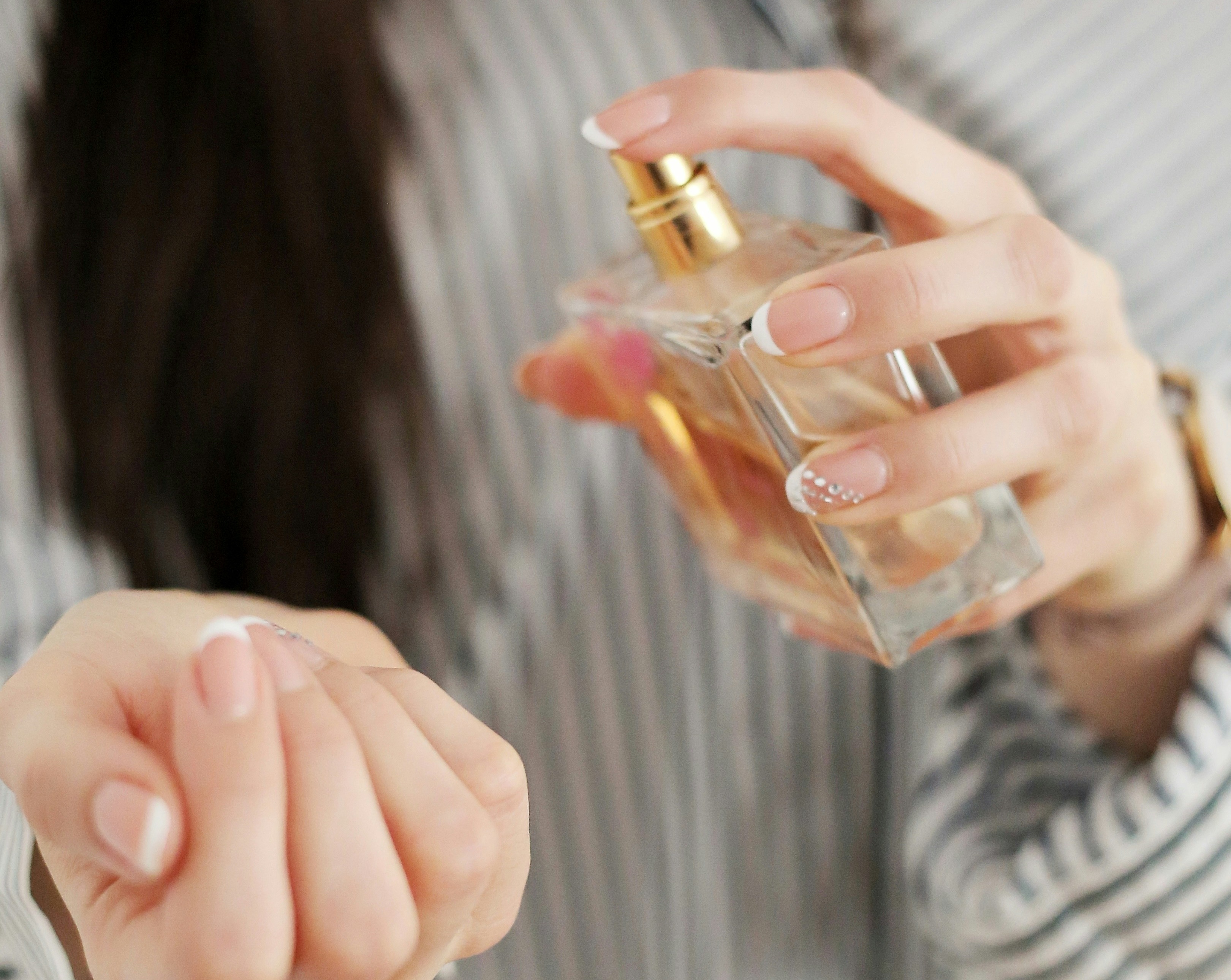 Mãos femininas com unhas francesinhas, uma delas com detalhes brilhantes, borrifando perfume de um frasco dourado e transparente no pulso esquerdo