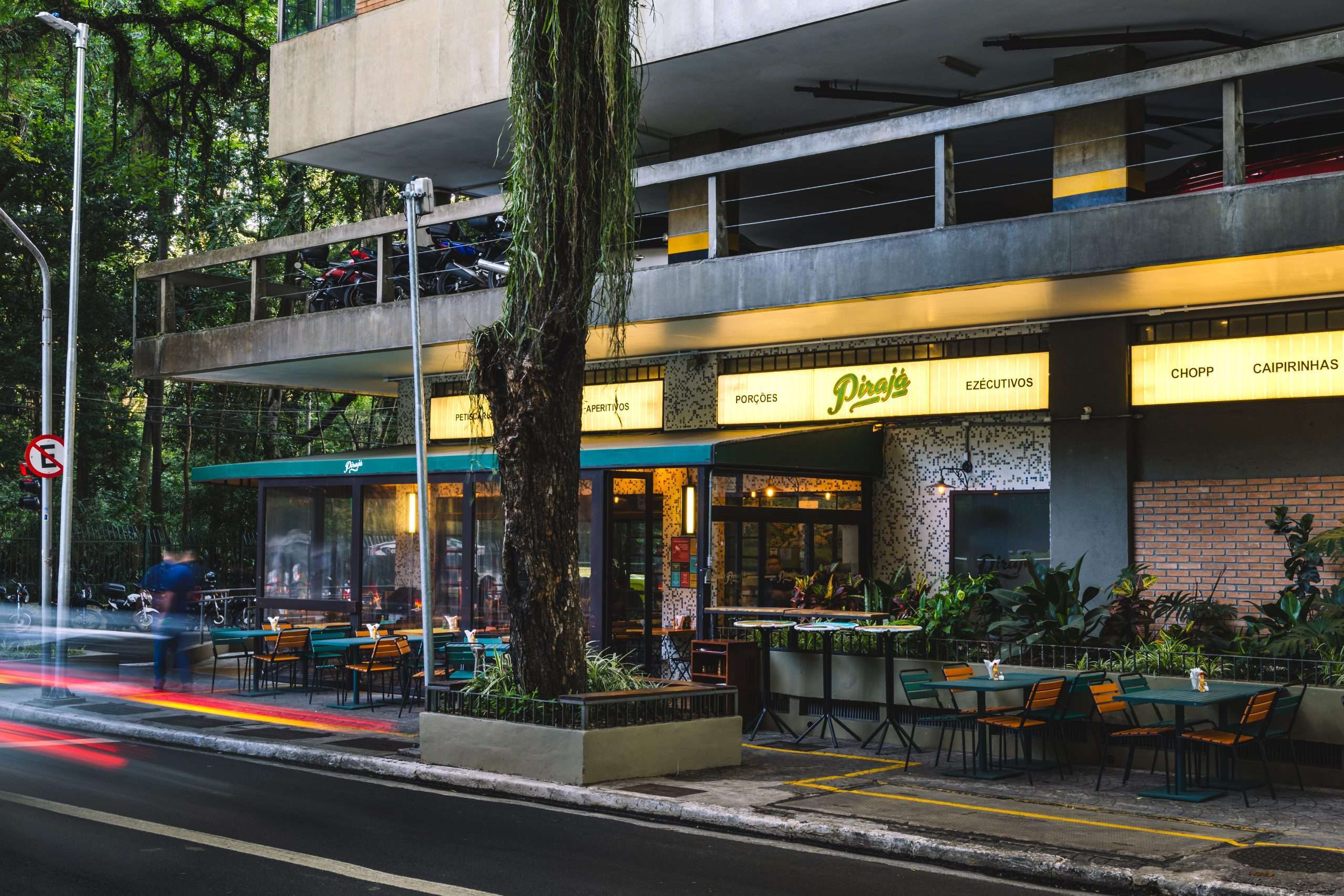 Fachada noturna do bar Piraj&aacute; com mesas e cadeiras na cal&ccedil;ada, sob um toldo verde. Placas iluminadas anunciam Petiscos, Aperitivos, Por&ccedil;&otilde;es, Executivos, Chopp e Caipirinhas. Um grande tronco de &aacute;rvore com musgo verde pende sobre a cal&ccedil;ada. No fundo, um pr&eacute;dio com estacionamento e &aacute;rvores. Rastros de luz vermelha e amarela de ve&iacute;culos em movimento na rua indicam longa exposi&ccedil;&atilde;o