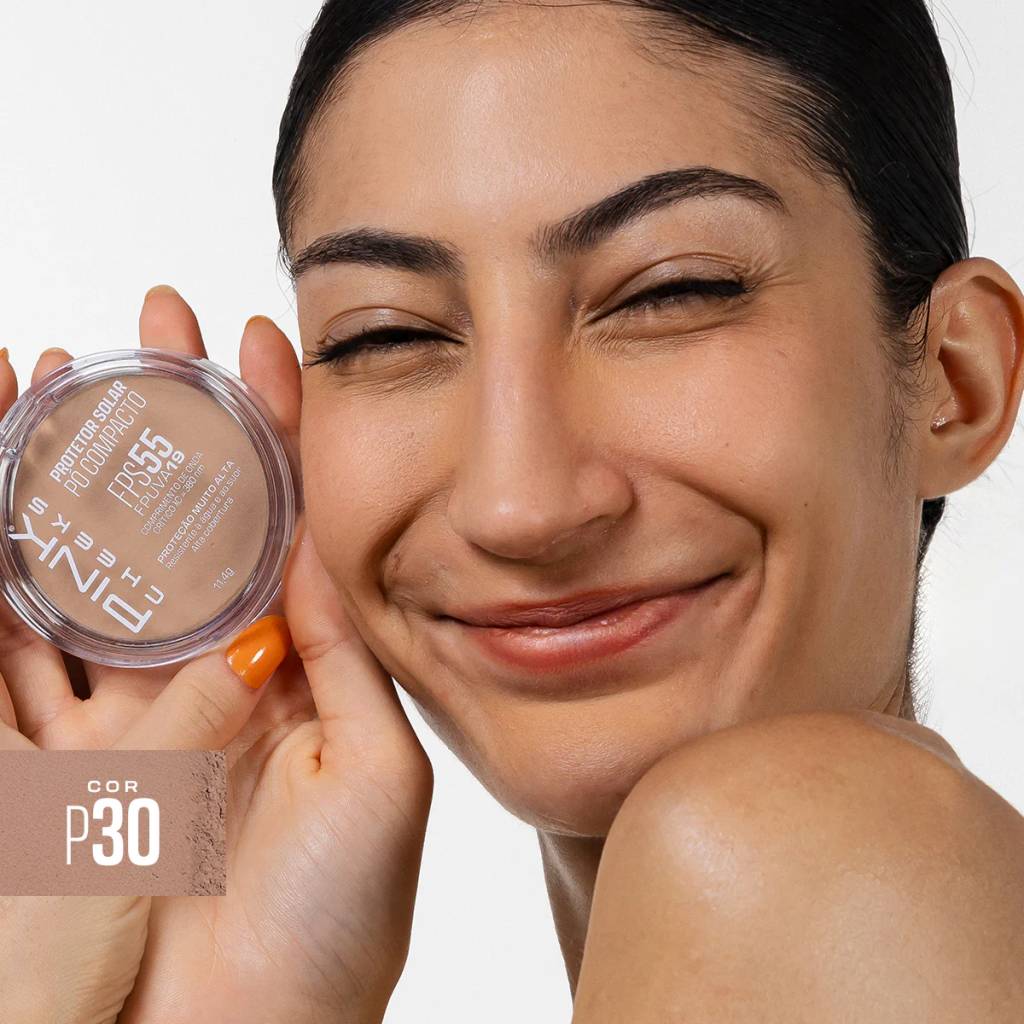 Mulher sorrindo com olhos fechados, segurando um p&oacute; compacto protetor solar da marca Pink Cheeks na cor P30. O produto tem FPS 55 e FPUVA 19, com prote&ccedil;&atilde;o muito alta e alta cobertura. Ela tem unhas laranjas e pele morena, com o cabelo preso e brincos discretos. O fundo &eacute; branco