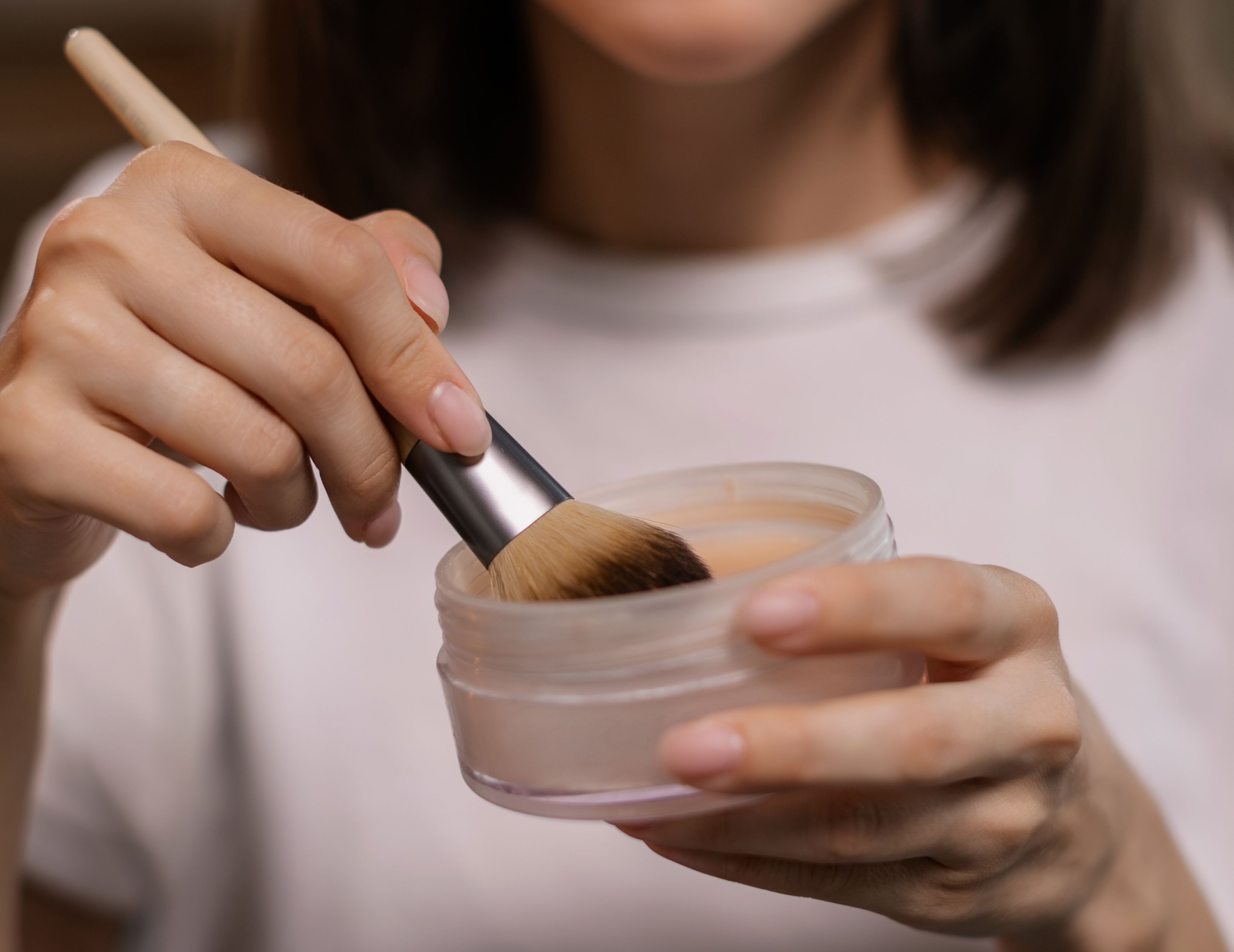 Mãos femininas segurando um pincel de maquiagem com cerdas marrons dentro de um pote transparente de pó facial solto