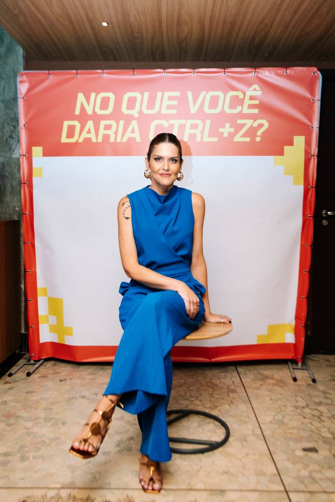 Mulher de cabelo preso, blusa azul sem mangas e cal&ccedil;a azul, sentada em um banco de madeira, sorrindo para a c&acirc;mera. Ao fundo, um banner vermelho com a frase NO QUE VOC&Ecirc; DARIA CTRL+Z? em letras amarelas e brancas