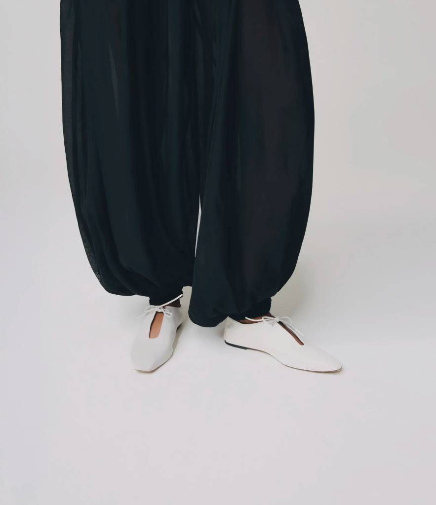 Pernas de uma pessoa vestindo cal&ccedil;as pretas largas e sapatos brancos de bico fino com cadar&ccedil;os brancos amarrados, em um fundo branco