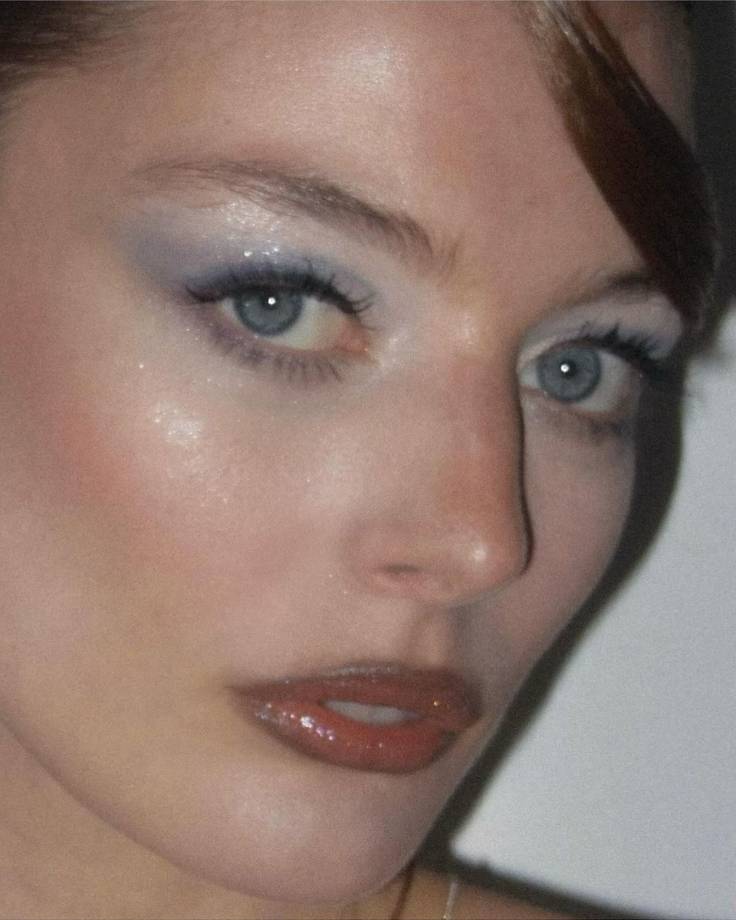 Rosto de mulher com maquiagem marcante, olhos azuis com sombra cintilante em tons de azul claro e delineador preto, l&aacute;bios entreabertos com batom vermelho brilhante e gloss, pele iluminada