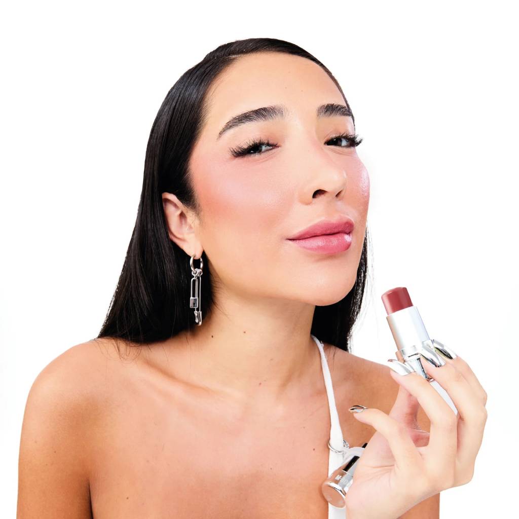 Mulher de cabelo preto liso, maquiagem rosa e brincos de alfinete, segurando um batom marrom avermelhado, sorrindo levemente para a c&acirc;mera
