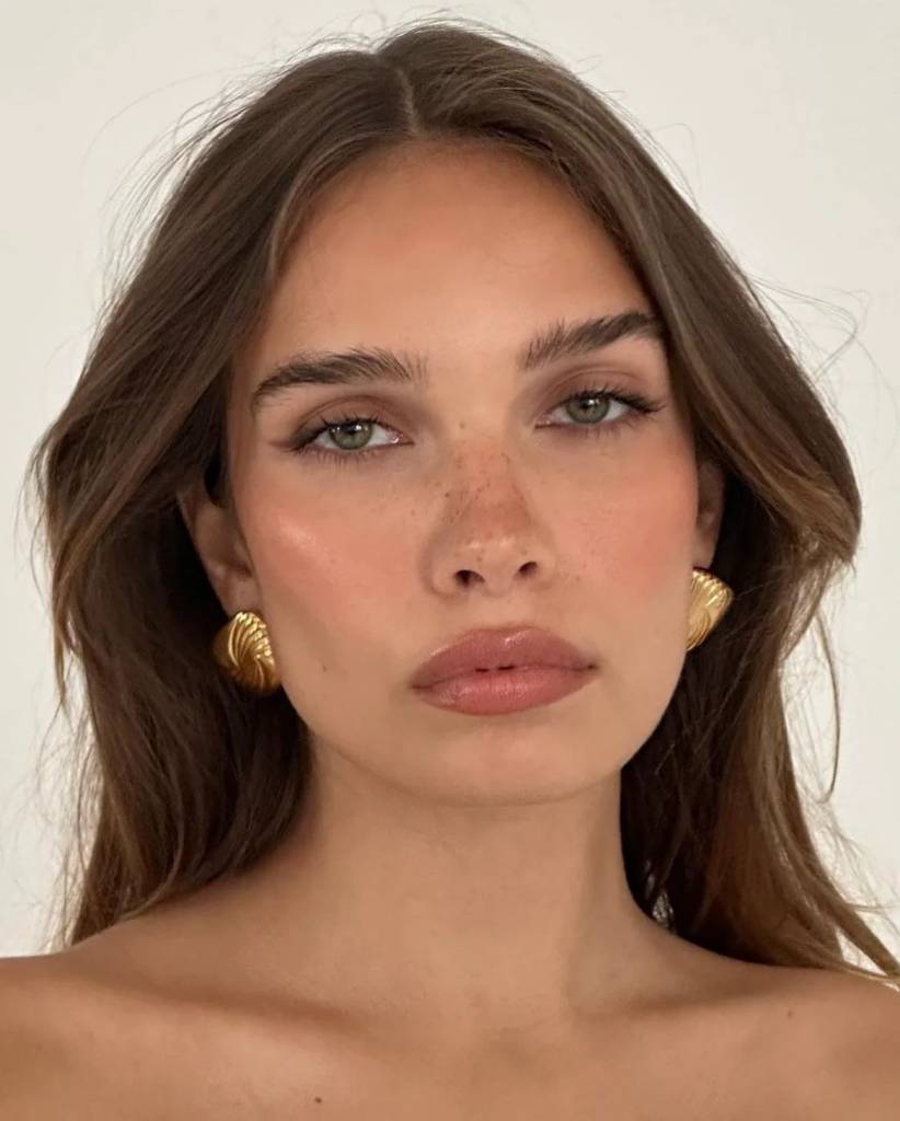Rosto de uma mulher jovem com cabelos castanhos soltos, olhos verdes, sardas no nariz e bochechas rosadas. Ela usa brincos dourados em formato de concha e batom nude brilhante.