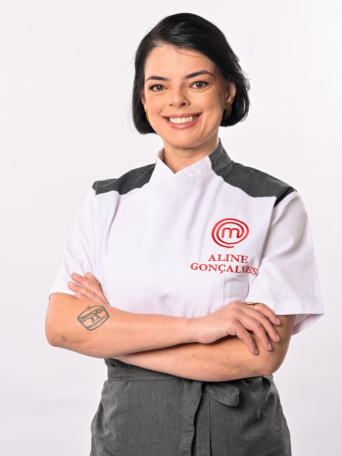 Mulher de pele clara, cabelo preto curto e liso, sorrindo, vestindo d&oacute;lm&atilde; branco com detalhes cinza nos ombros e avental cinza. O d&oacute;lm&atilde; tem o logo vermelho do MasterChef e o nome Aline Gon&ccedil;alves. Ela tem uma tatuagem de bolo no antebra&ccedil;o esquerdo e brincos amarelos. Os bra&ccedil;os est&atilde;o cruzados na altura da cintura, e ela usa um anel dourado no dedo anelar direito