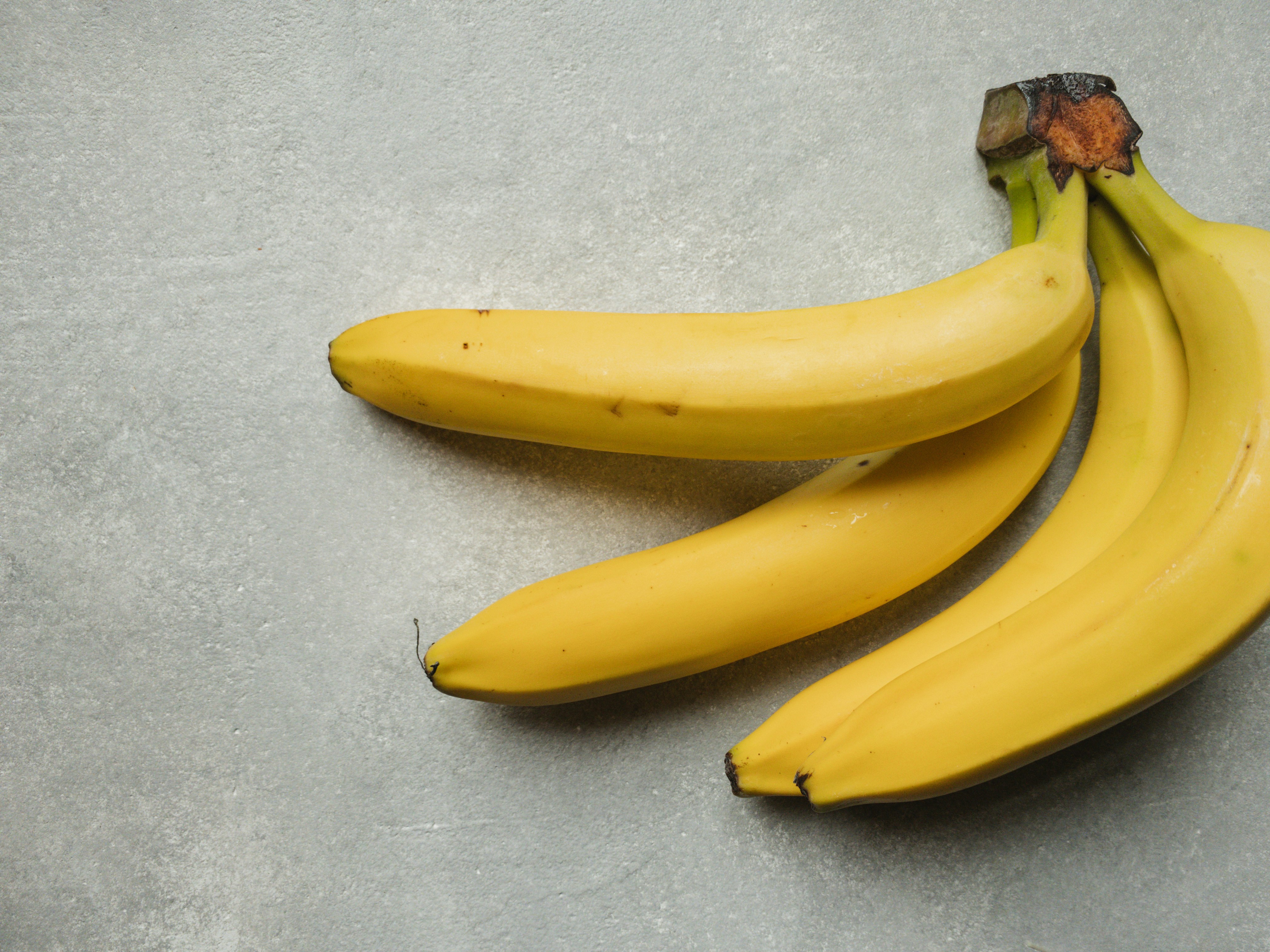 Tr&ecirc;s bananas maduras, de casca amarela vibrante com pequenas manchas marrons, dispostas sobre uma superf&iacute;cie cinza texturizada